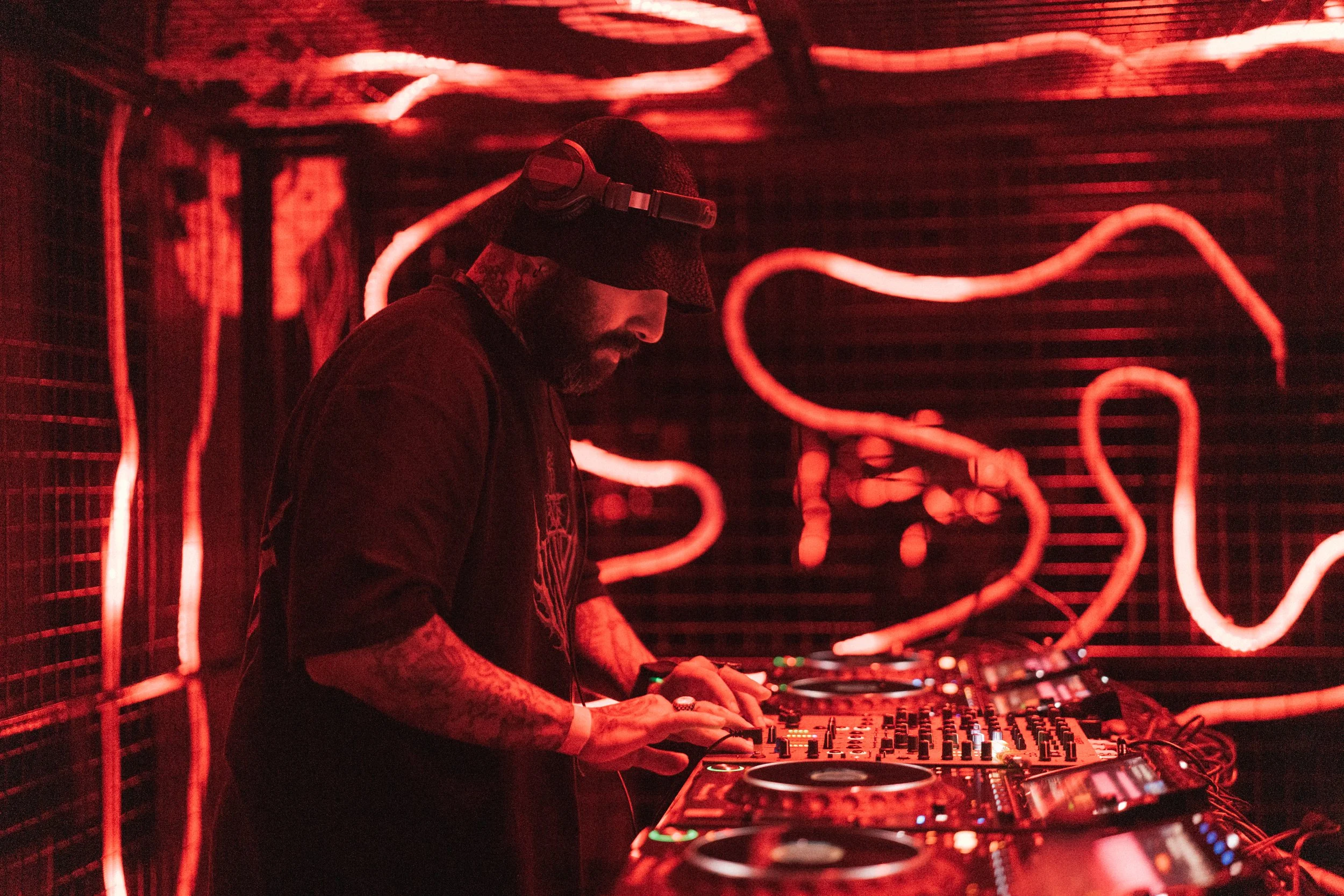 A DJ wearing headphones and a cap, mixing music on a DJ console in a dimly lit room with red neon lights on the wall.