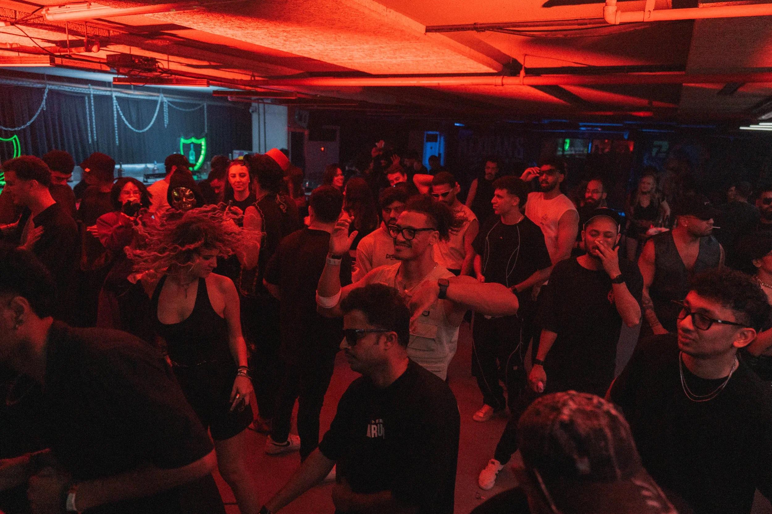 People dancing and socializing in a dark nightclub with red and blue lighting, neon shield, and sword decorations.