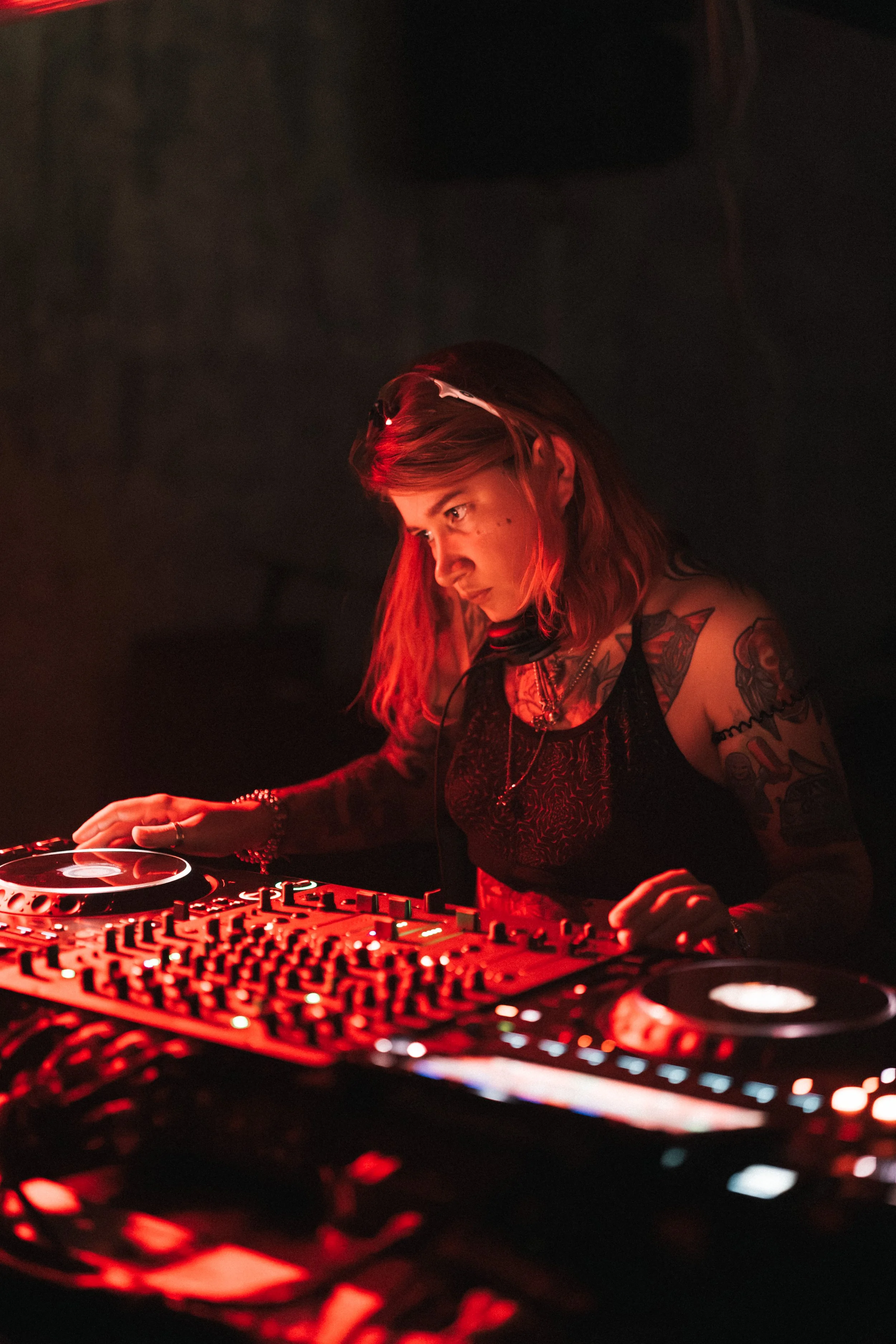 A female DJ with red hair, tattoos, and a black lace top DJing at a club with red lighting.