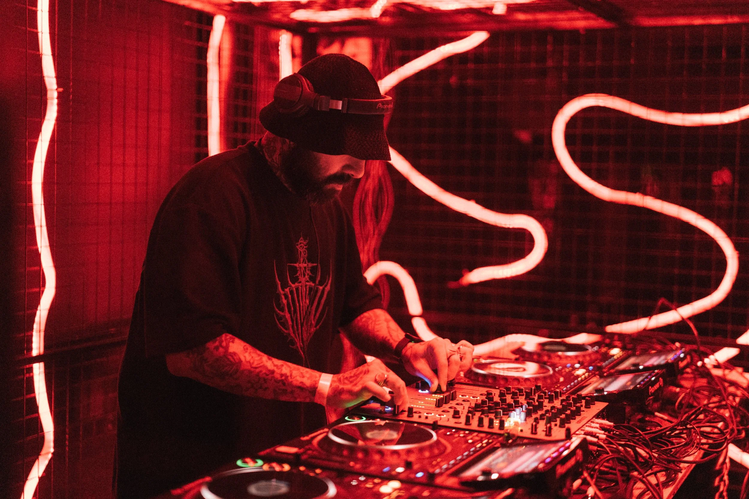 A DJ wearing a black shirt with a design, a black hat, and headphones, mixing music on DJ equipment in a red-lit venue with neon light decorations on the wall.