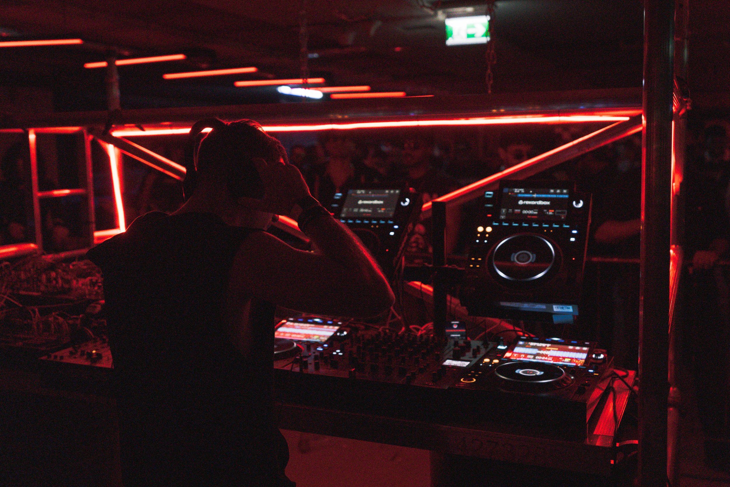A DJ performing at a nightclub with red neon lighting, using a mixing console and headphones.