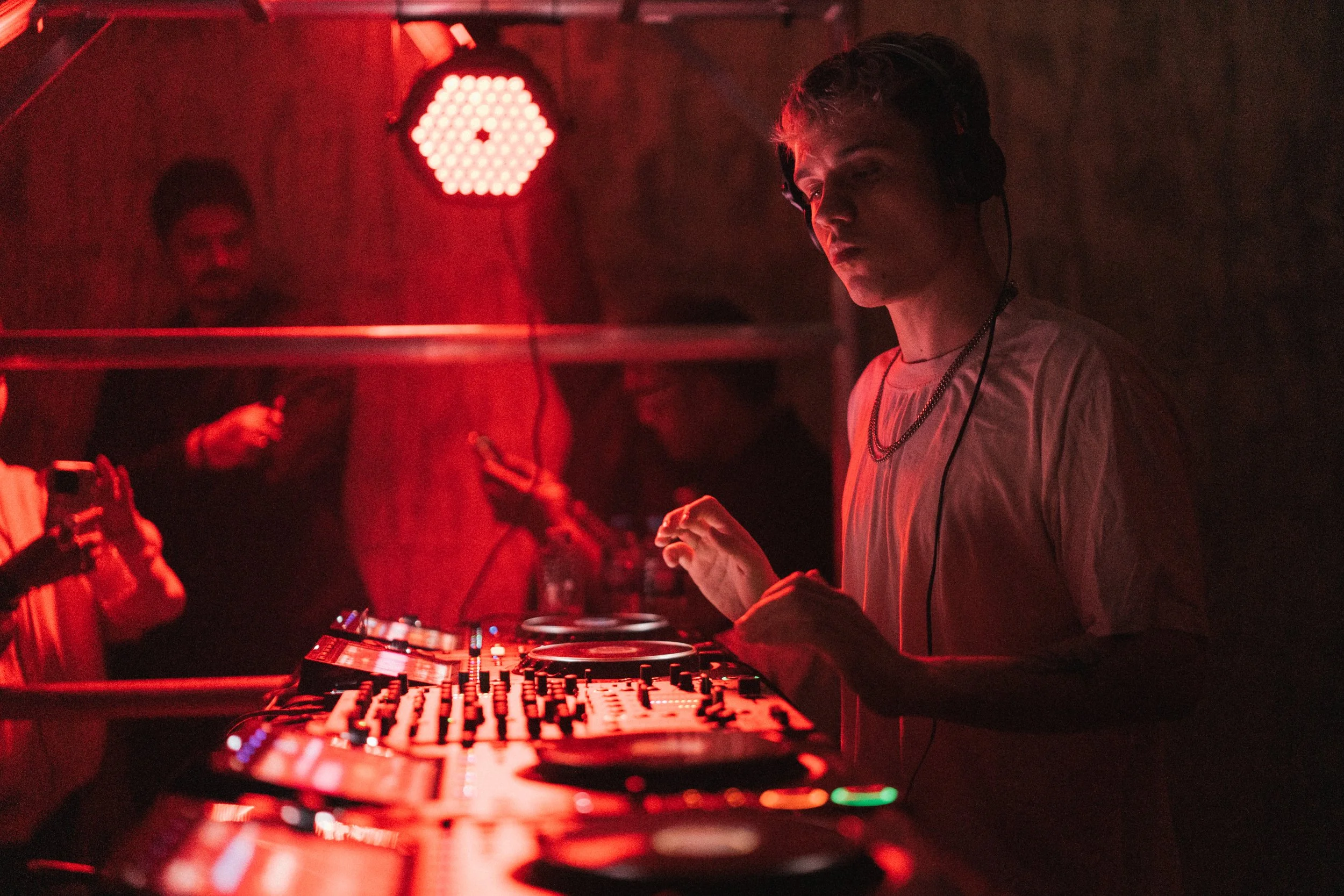A DJ with headphones playing music at a nightclub with red lighting, while people in the background are engaged with their phones.