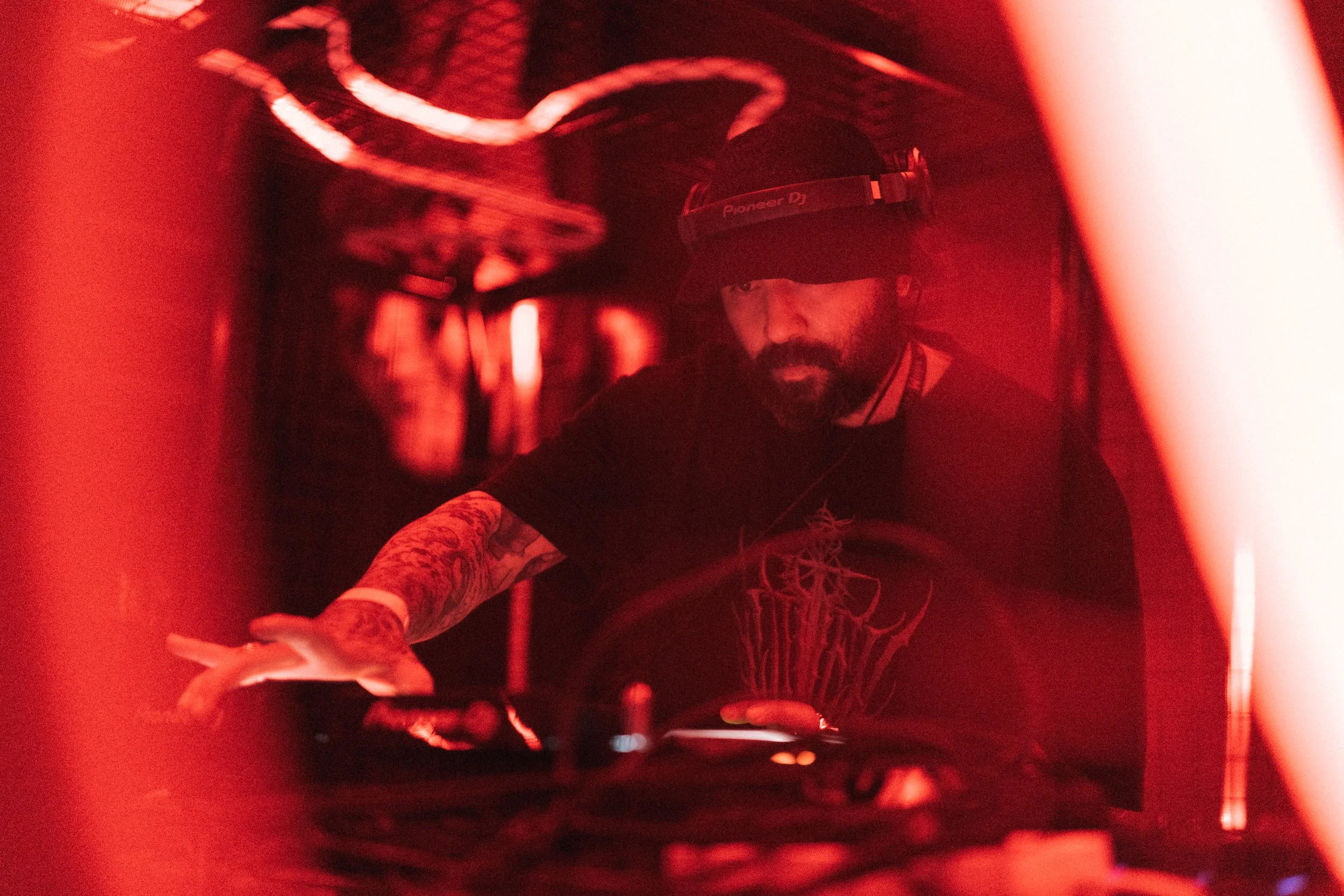 A DJ with tattoos, a beard, and wearing a Pioneer DJ headband, mixing music in a dimly lit environment with red lighting and blurred background of people.