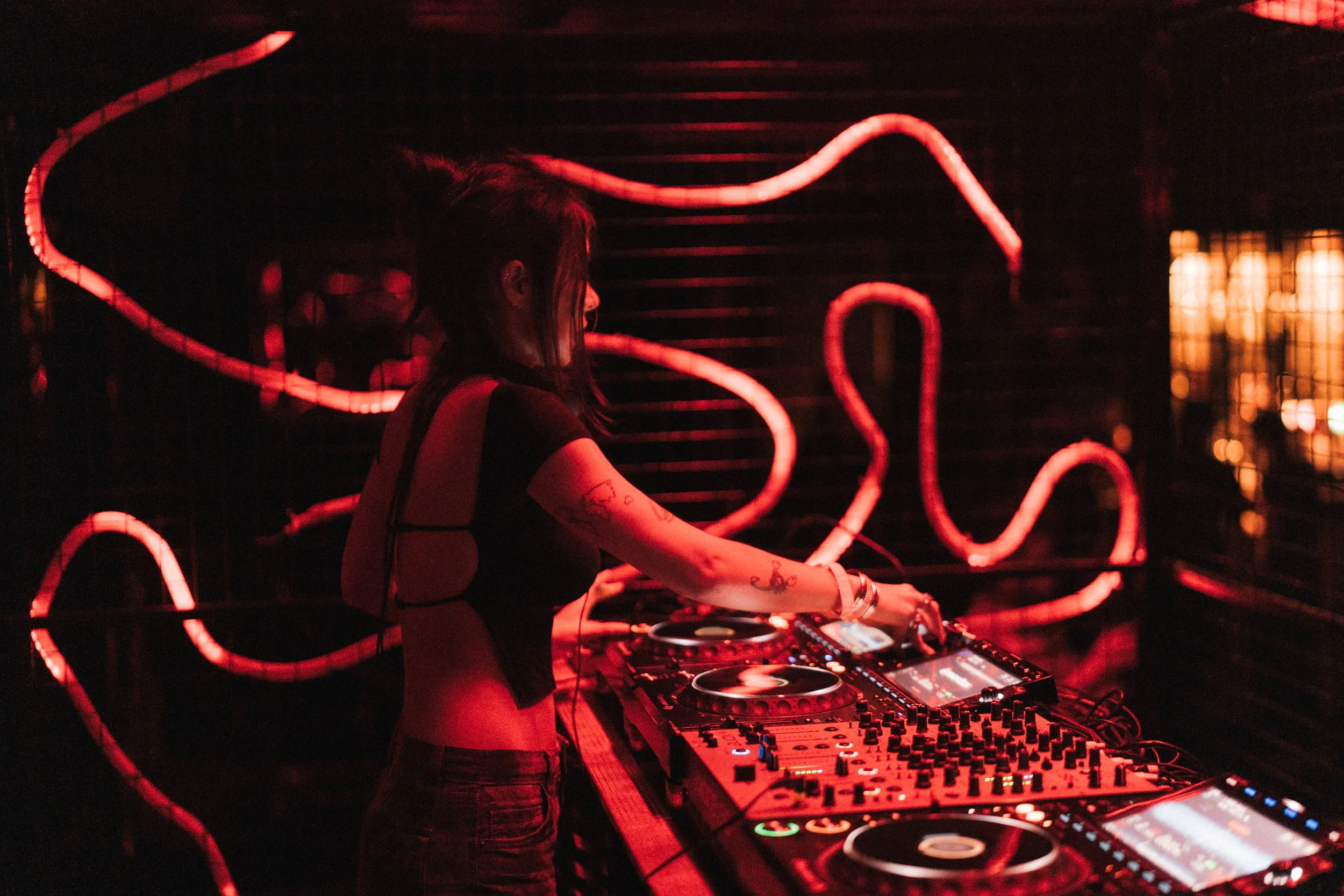 A female DJ is performing at a dark nightclub illuminated by red neon lights on the wall behind her. She is mixing music on DJ equipment, wearing a black top, with tattoos on her arms.