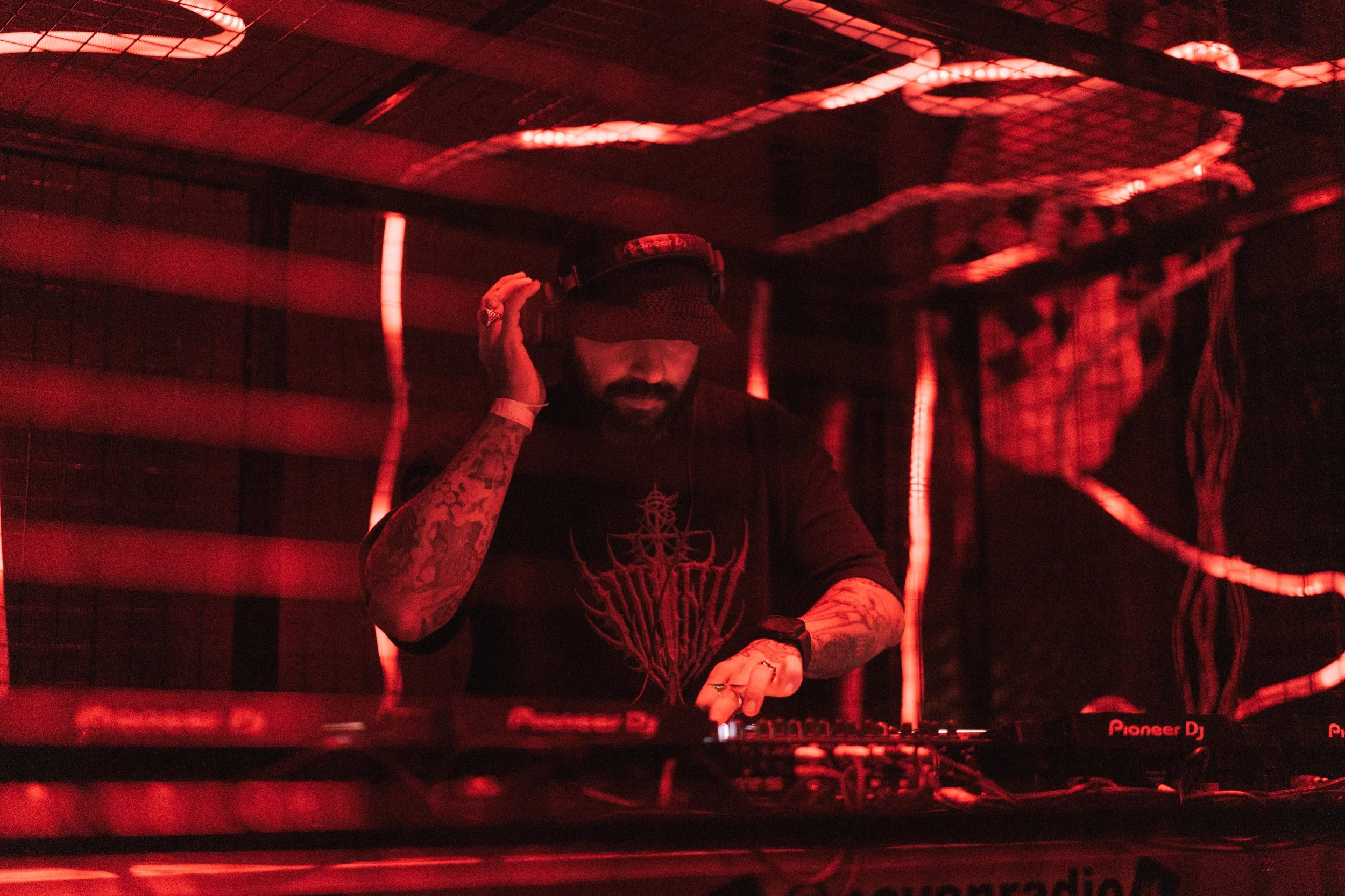 A DJ with tattoos on his arms, wearing a black cap and shirt, is performing in a dimly lit space with red lighting, operating DJ equipment behind a metal barrier.