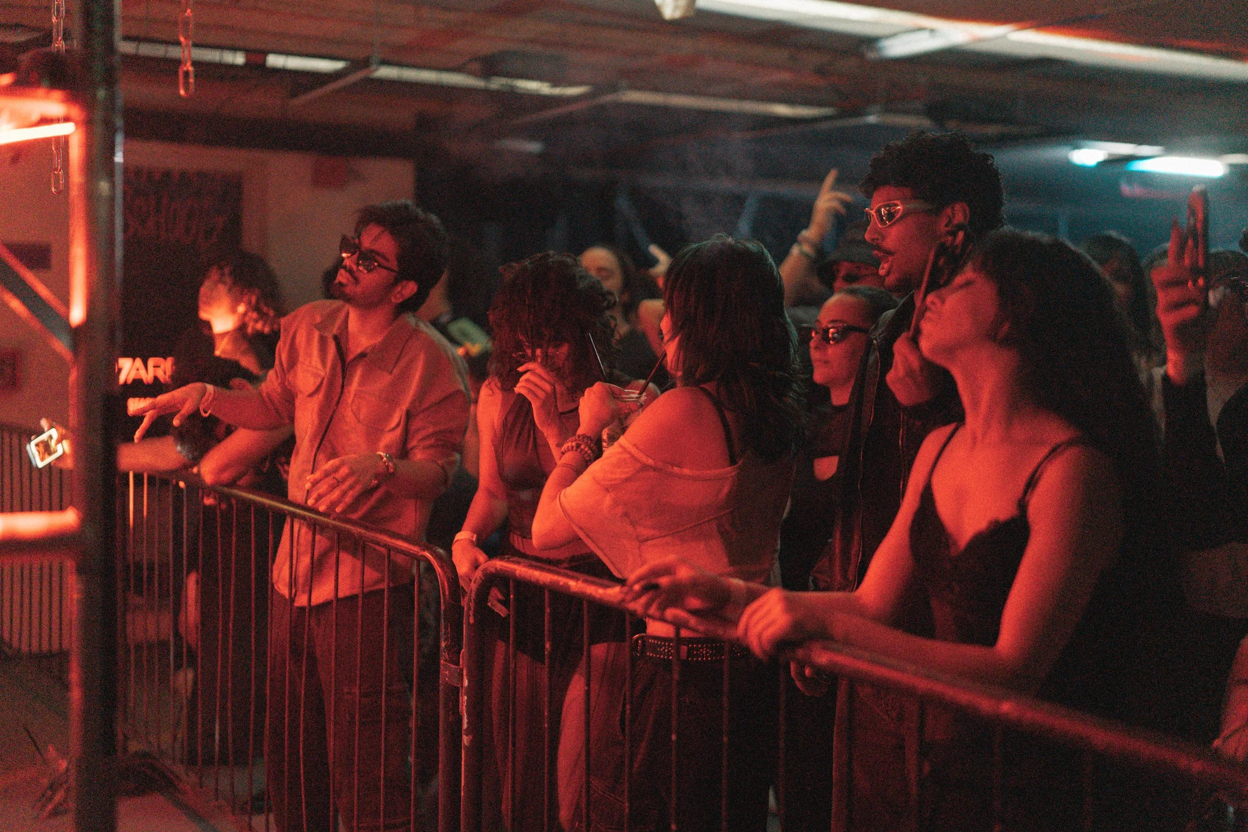 People at a music concert or event standing behind a metal barricade, illuminated by red and blue stage lighting.