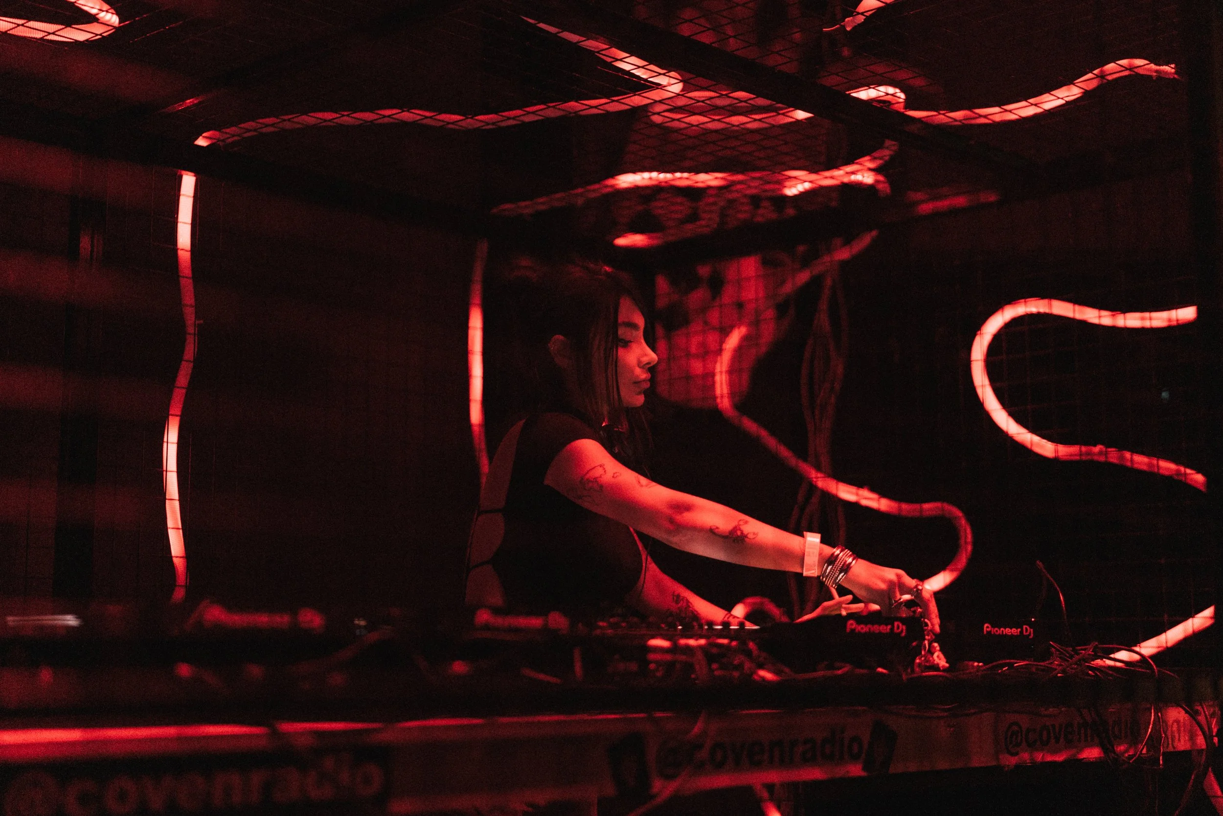 A female DJ performing in a dark club with red lighting and neon snake designs on the wall behind her.