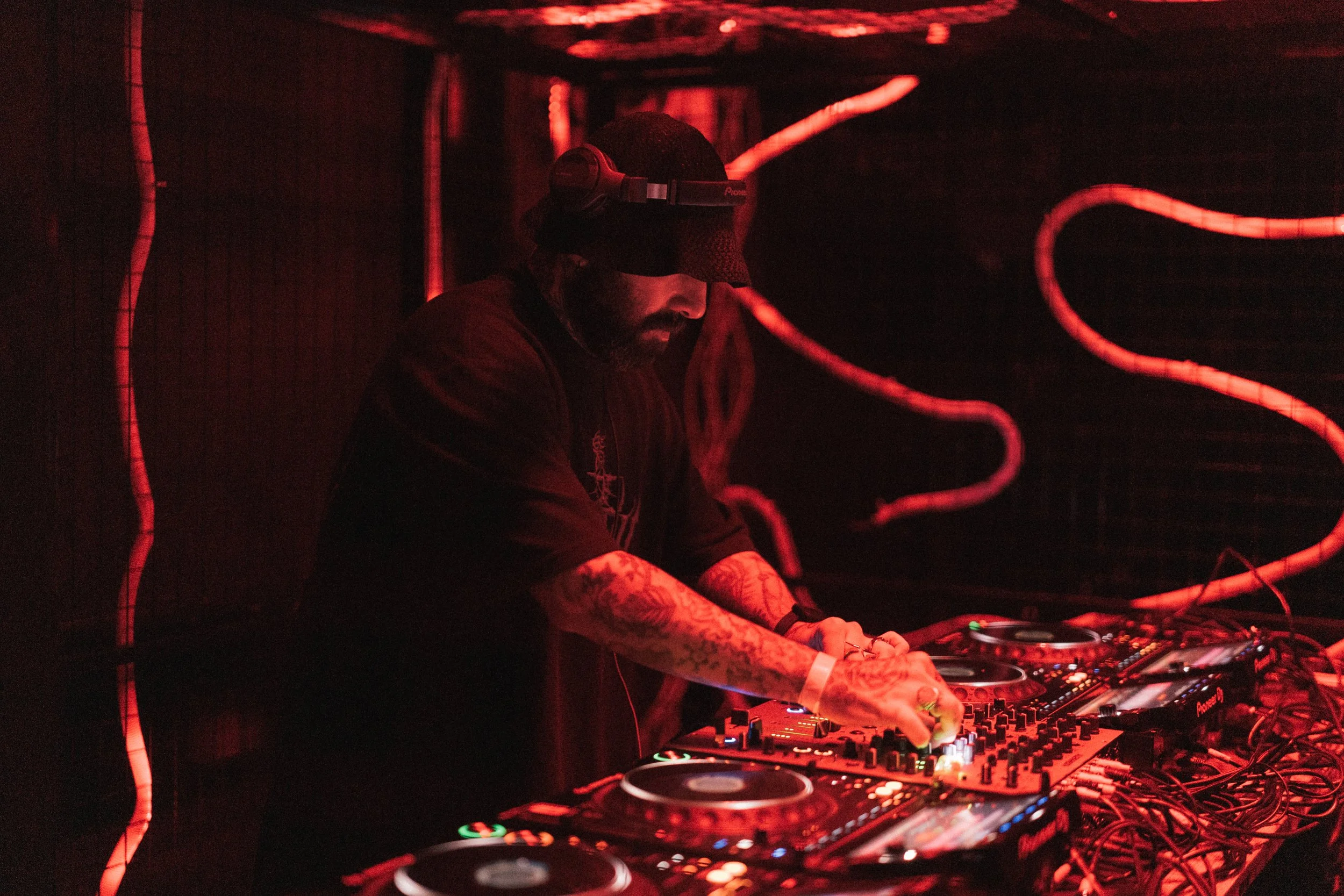 A DJ wearing headphones and a black beanie is performing at a dark nightclub with red lighting, using professional DJ equipment including turntables and a mixer.