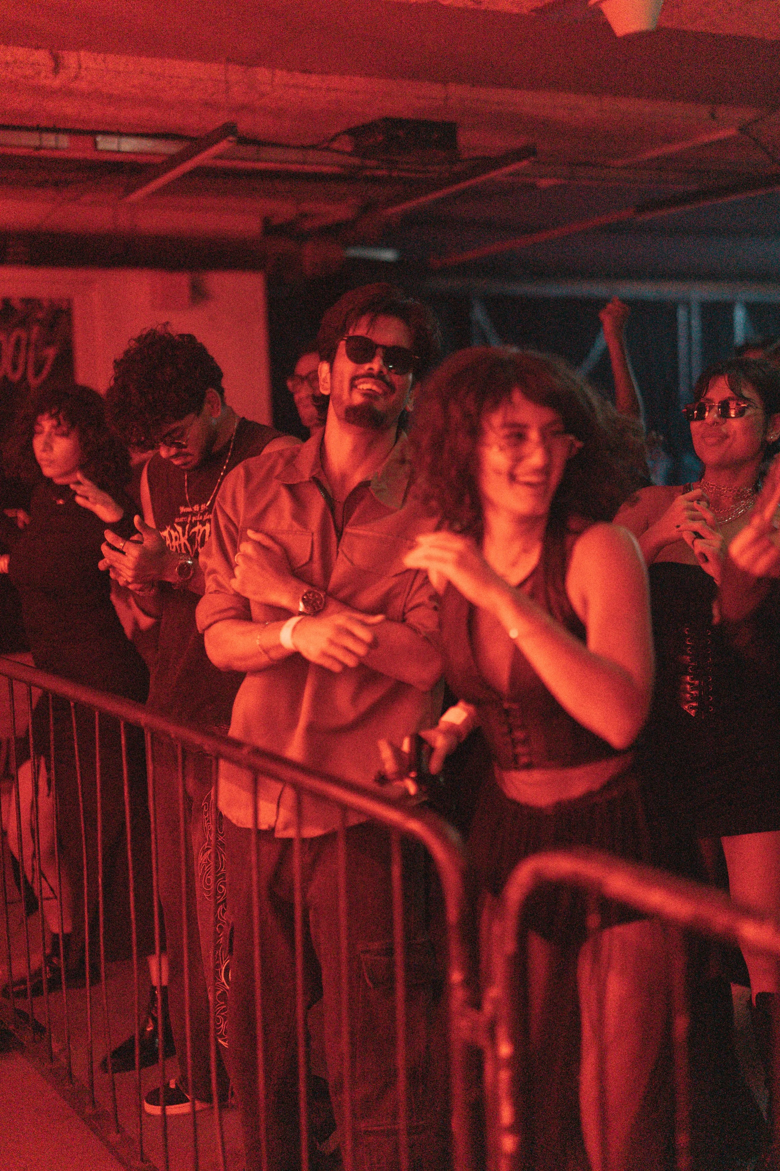 People enjoying a concert or event behind a metal barricade, some wearing sunglasses and casual clothing, with a red lighting glow.