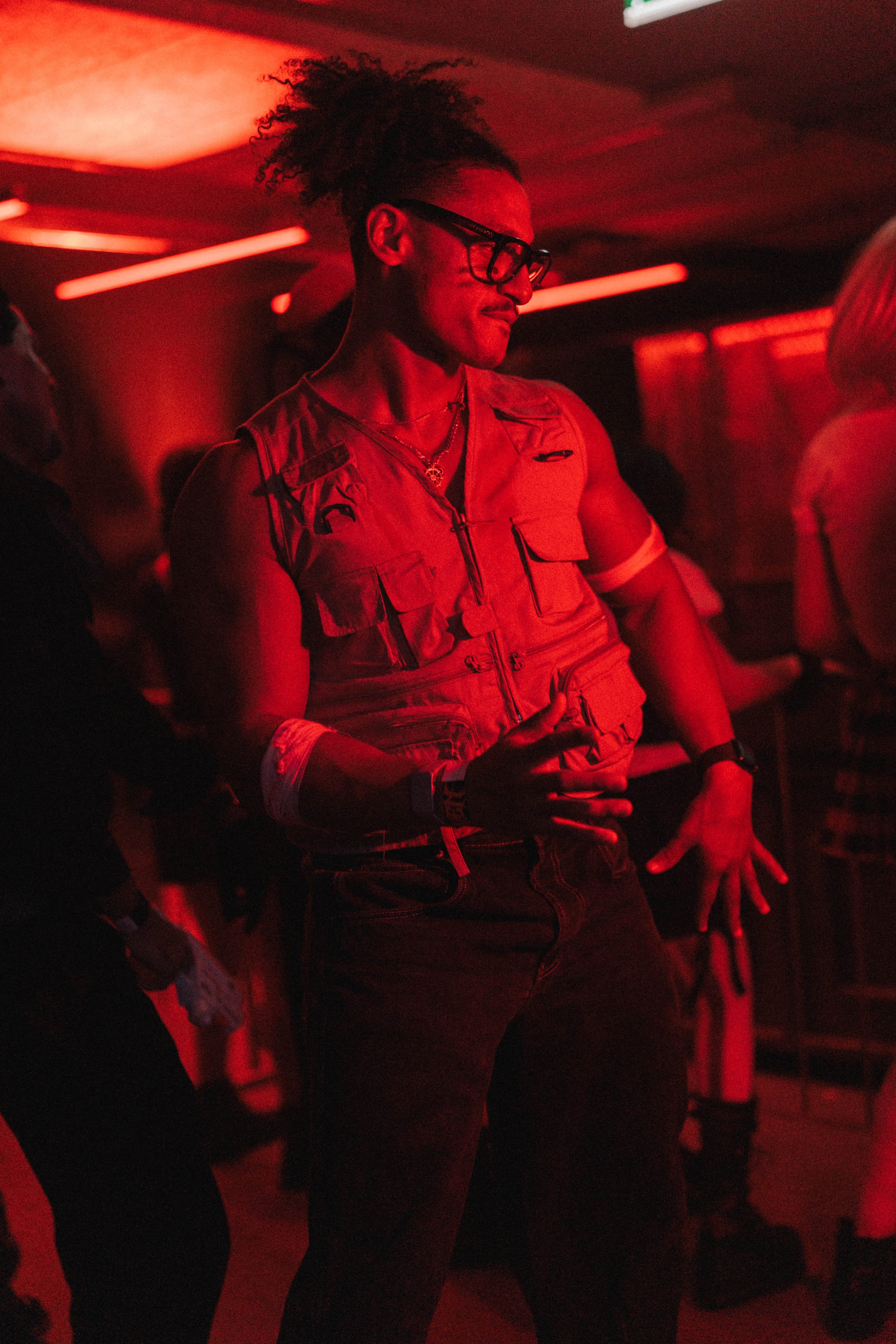 A person with curly hair in a ponytail, wearing glasses and a sleeveless vest dancing in a red-lit environment.