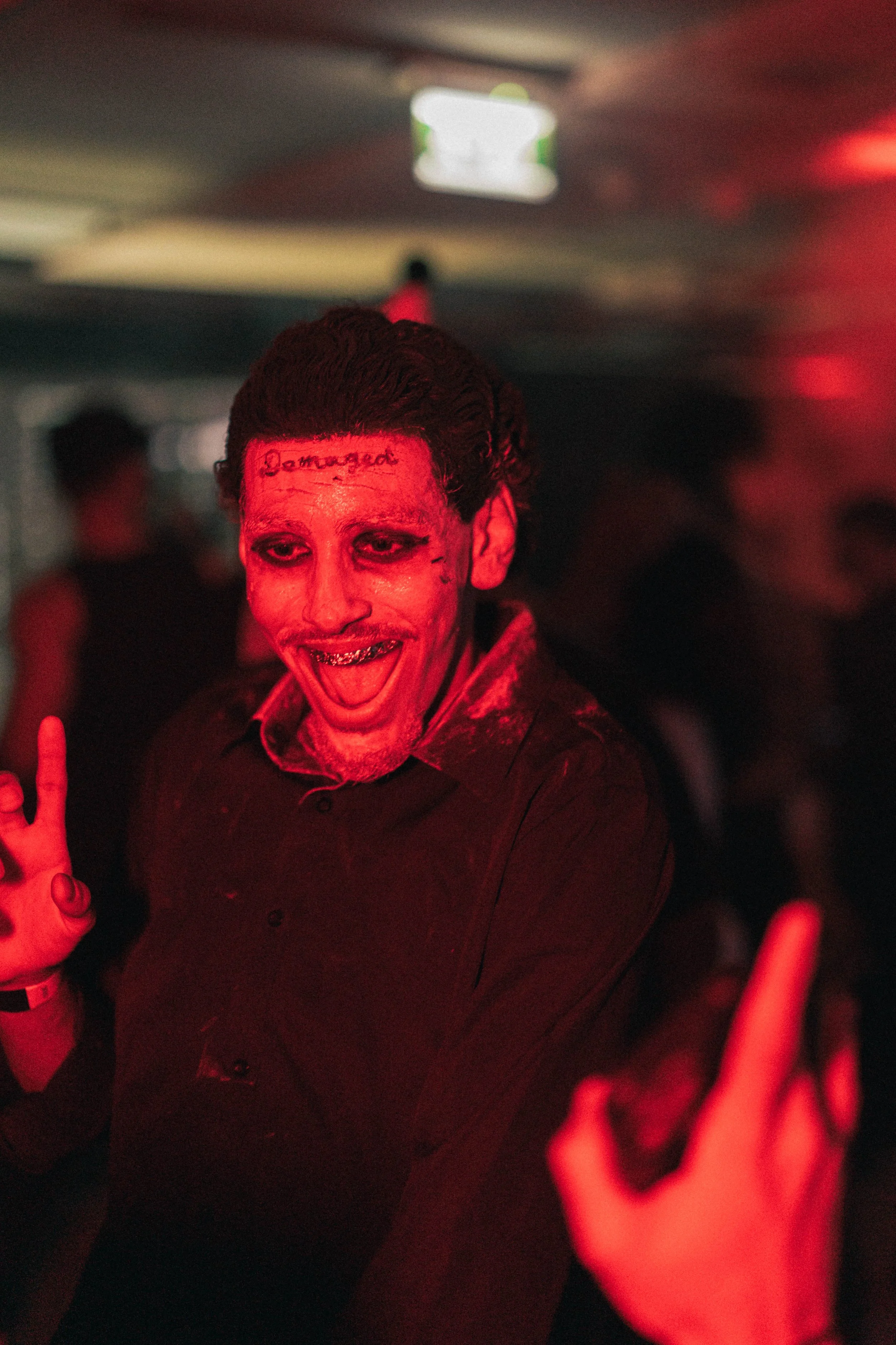 A person dressed up as a scary clown with face makeup, wearing a black shirt, smiling and making a gesture with one hand at a party or event with red lighting.