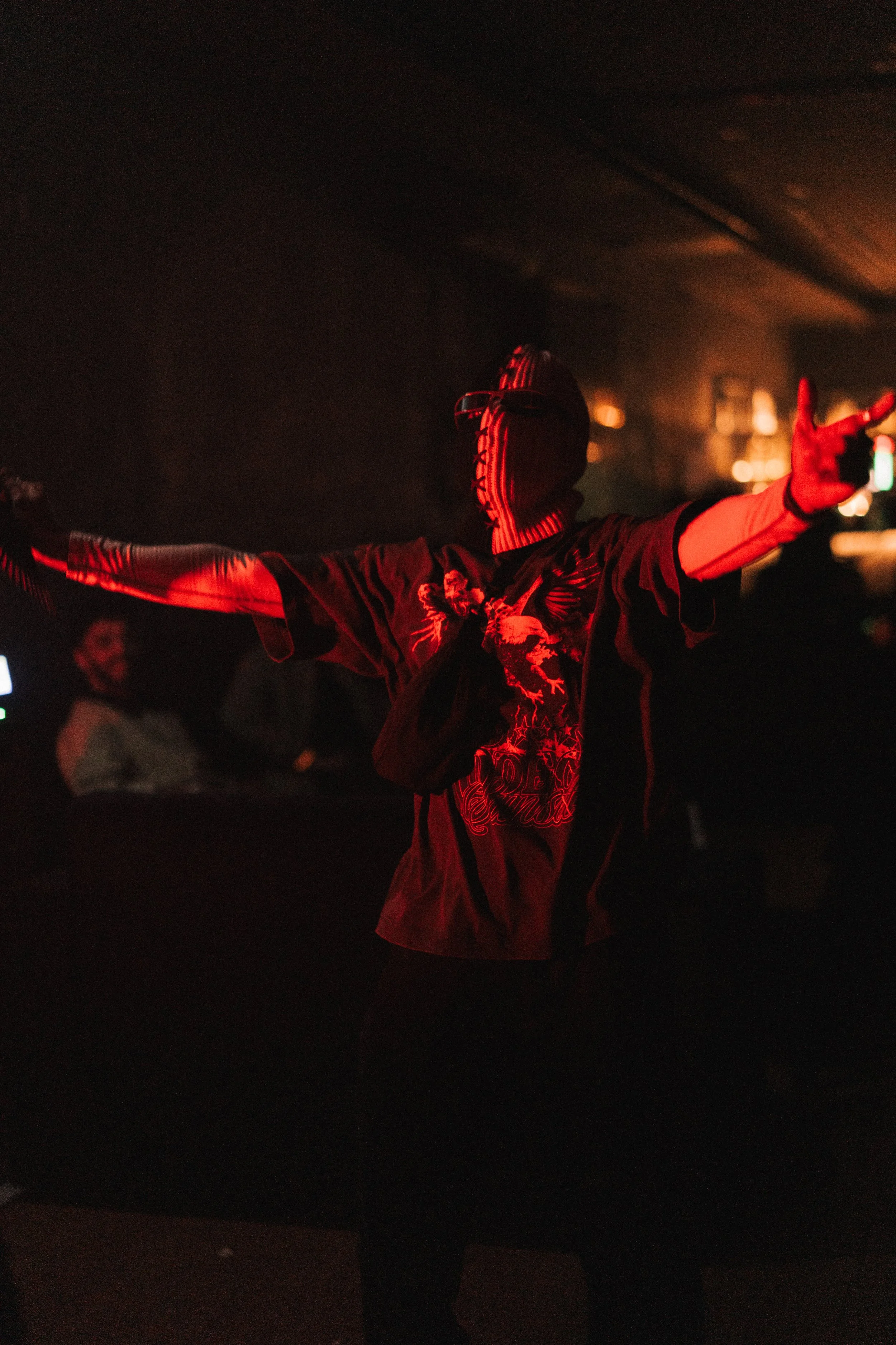 Person with arms outstretched in a dark room illuminated by red light, wearing a mask and a T-shirt with a design.