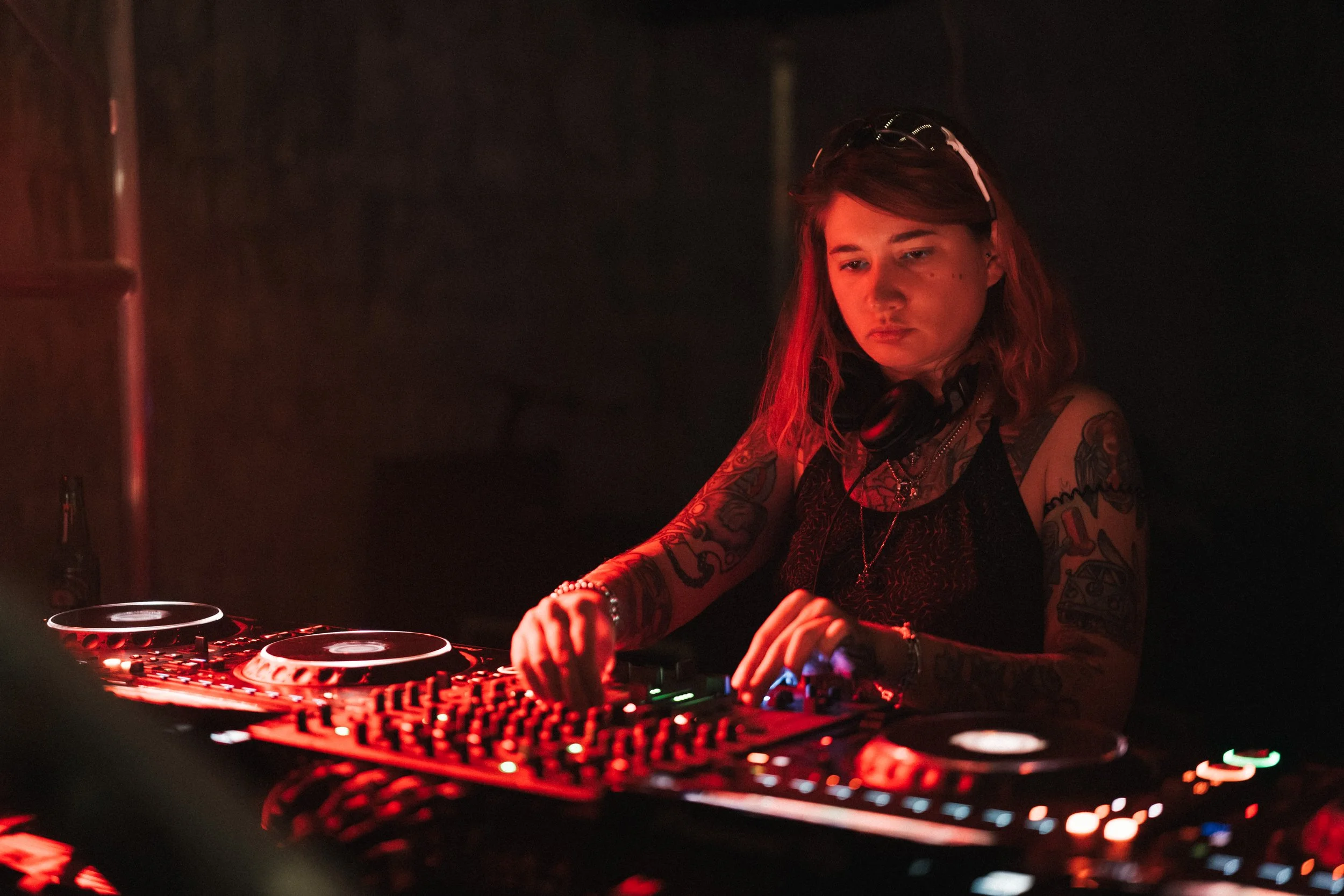 A young female DJ with tattoos on her arms, wearing headphones and a black top, operating a DJ mixer with glowing controls in a dark environment.