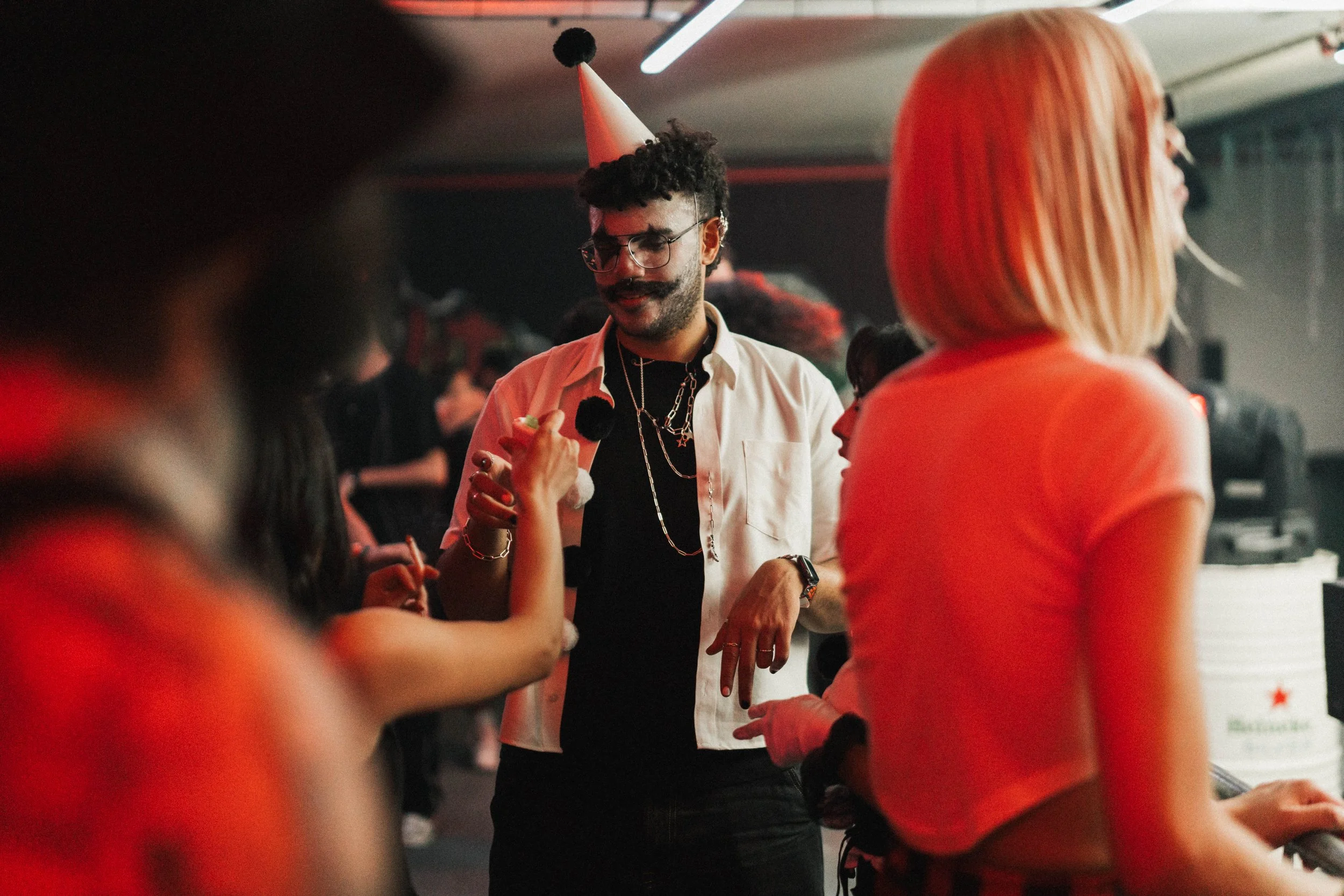 A man with glasses, a mustache, and curly hair wearing a white shirt, black T-shirt, and necklaces, holding a drink and wearing a party hat, is socializing at a party with other people in a dimly lit room.