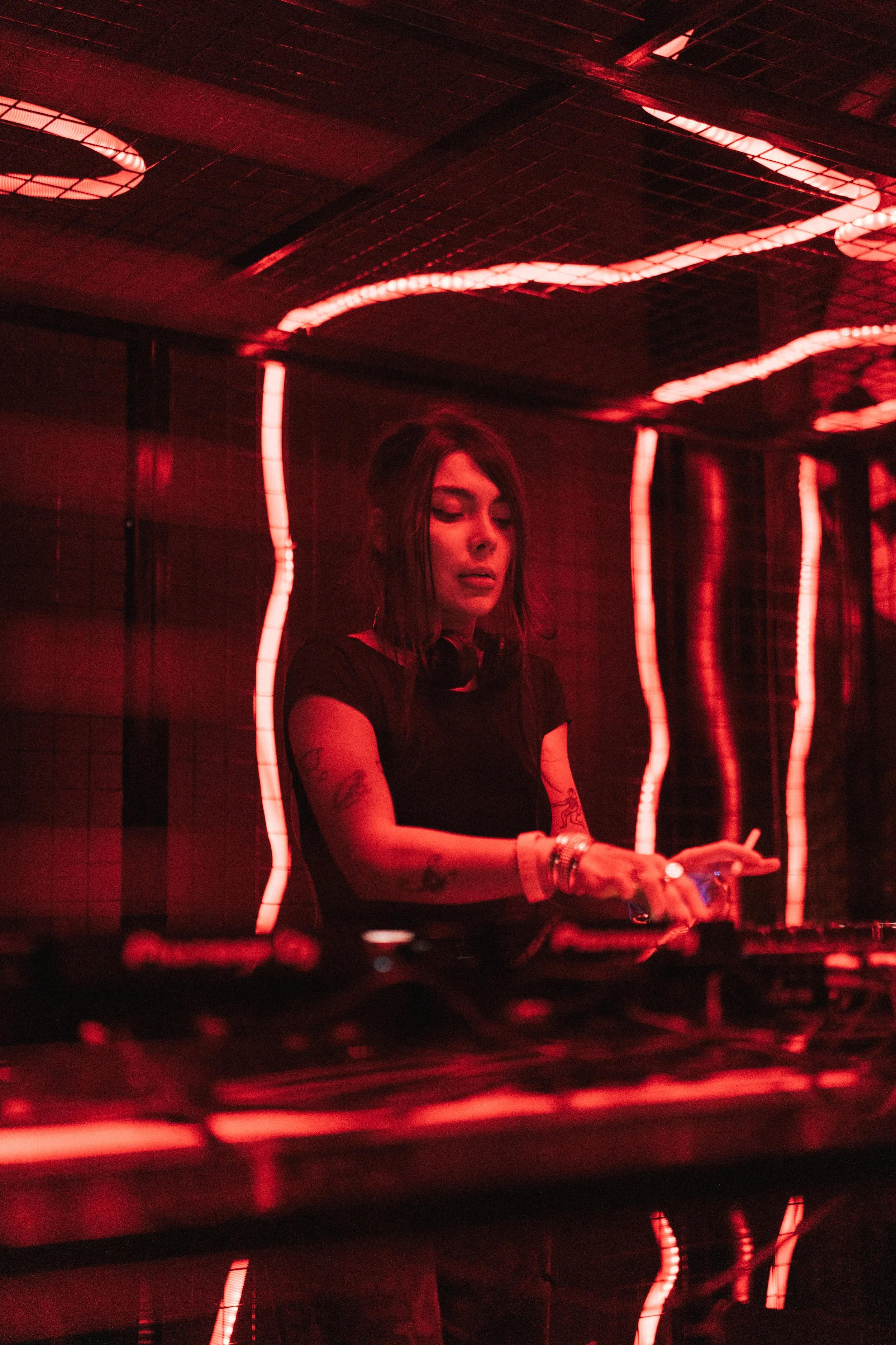 A female DJ with tattoos on her arms, wearing headphones around her neck, performing in a dimly lit club with red neon lights.