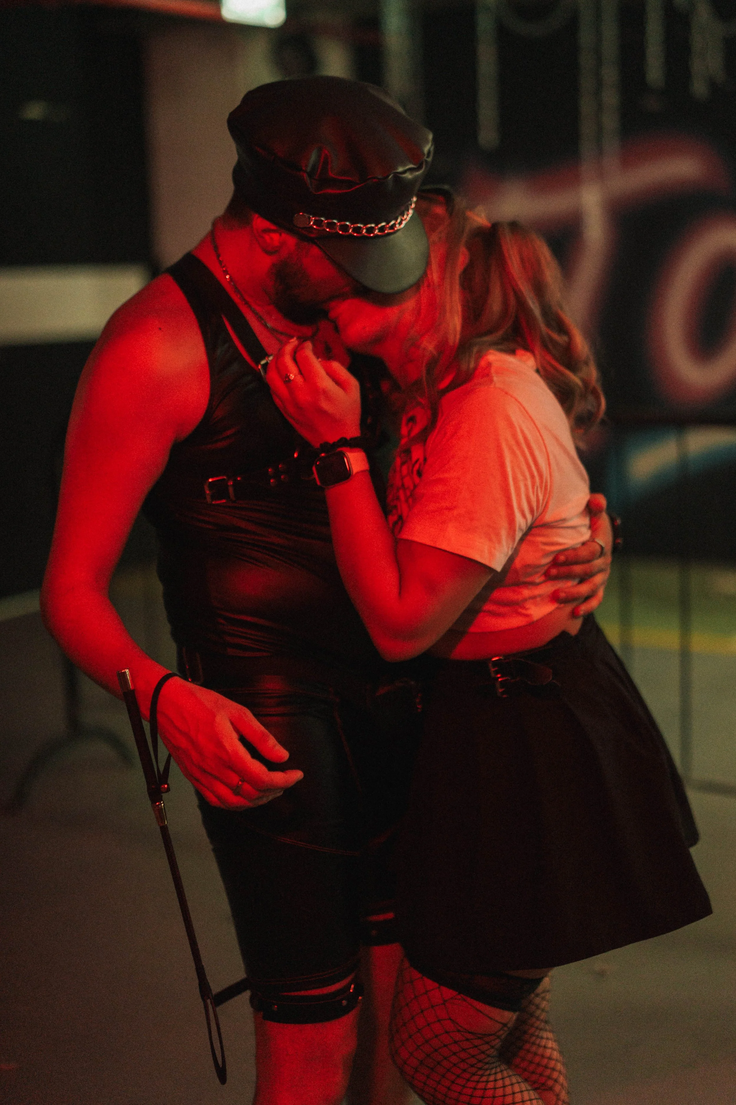 A man and woman in an intimate embrace, with the man wearing a leather cap and outfit, and the woman wearing a skirt, fishnet stockings, and a t-shirt under her unzipped jacket, sharing a kiss in a dimly lit room with a red hue.