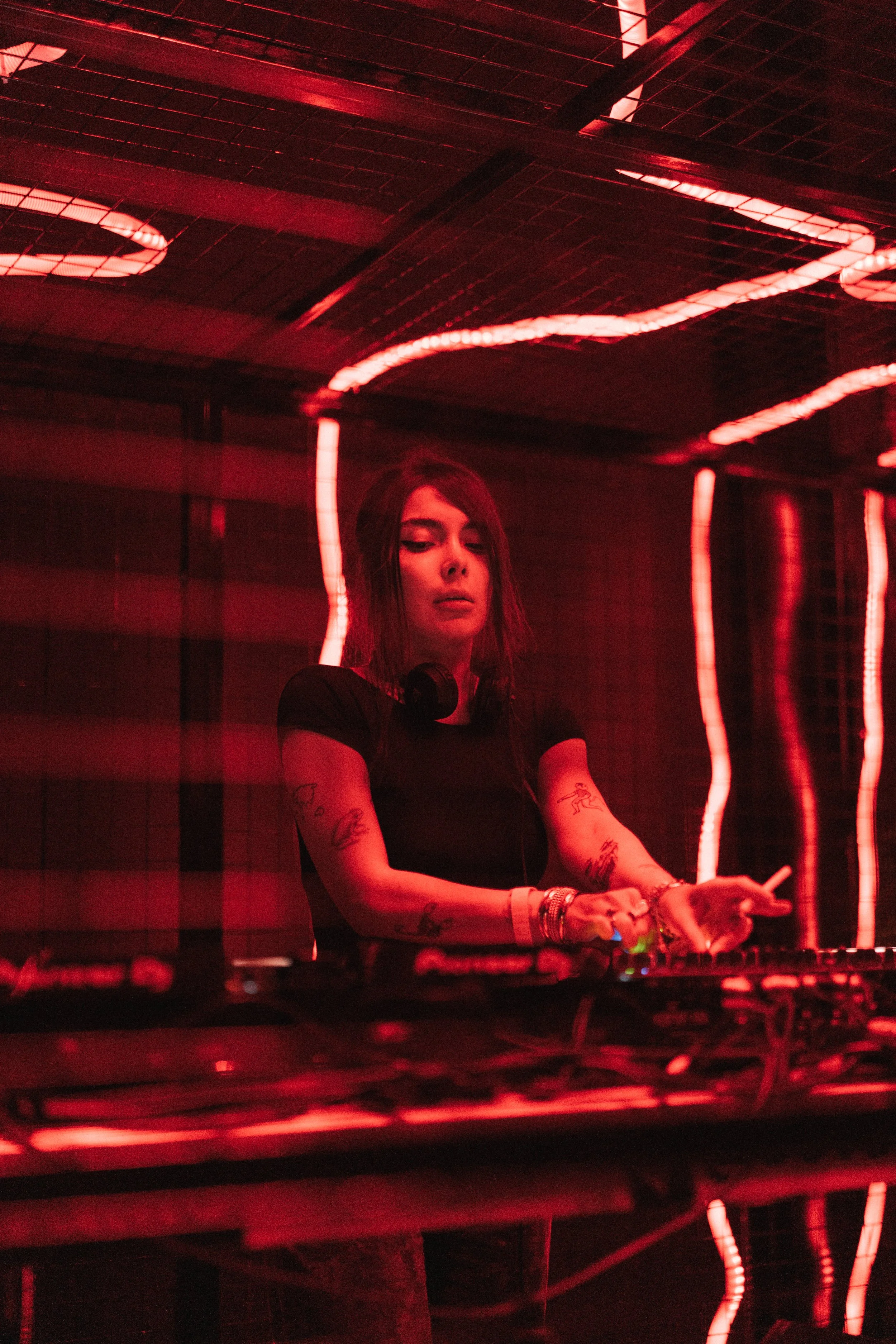 Female DJ with tattoos wearing black shirt, surrounded by red neon lights, operating DJ equipment in a dark setting.