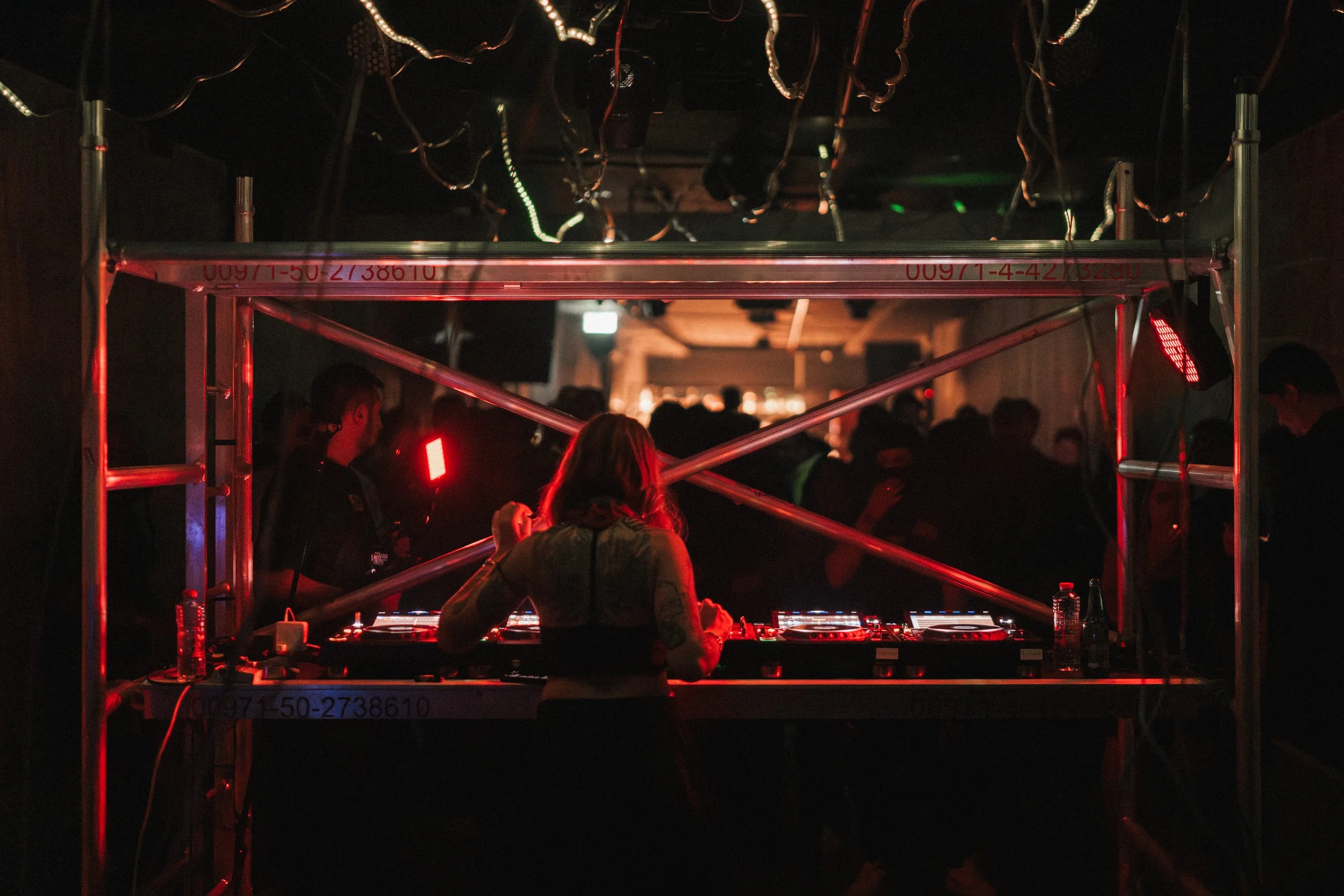 A DJ is performing at a nightclub, surrounded by a crowd of people dancing in a dark, red-lit environment.