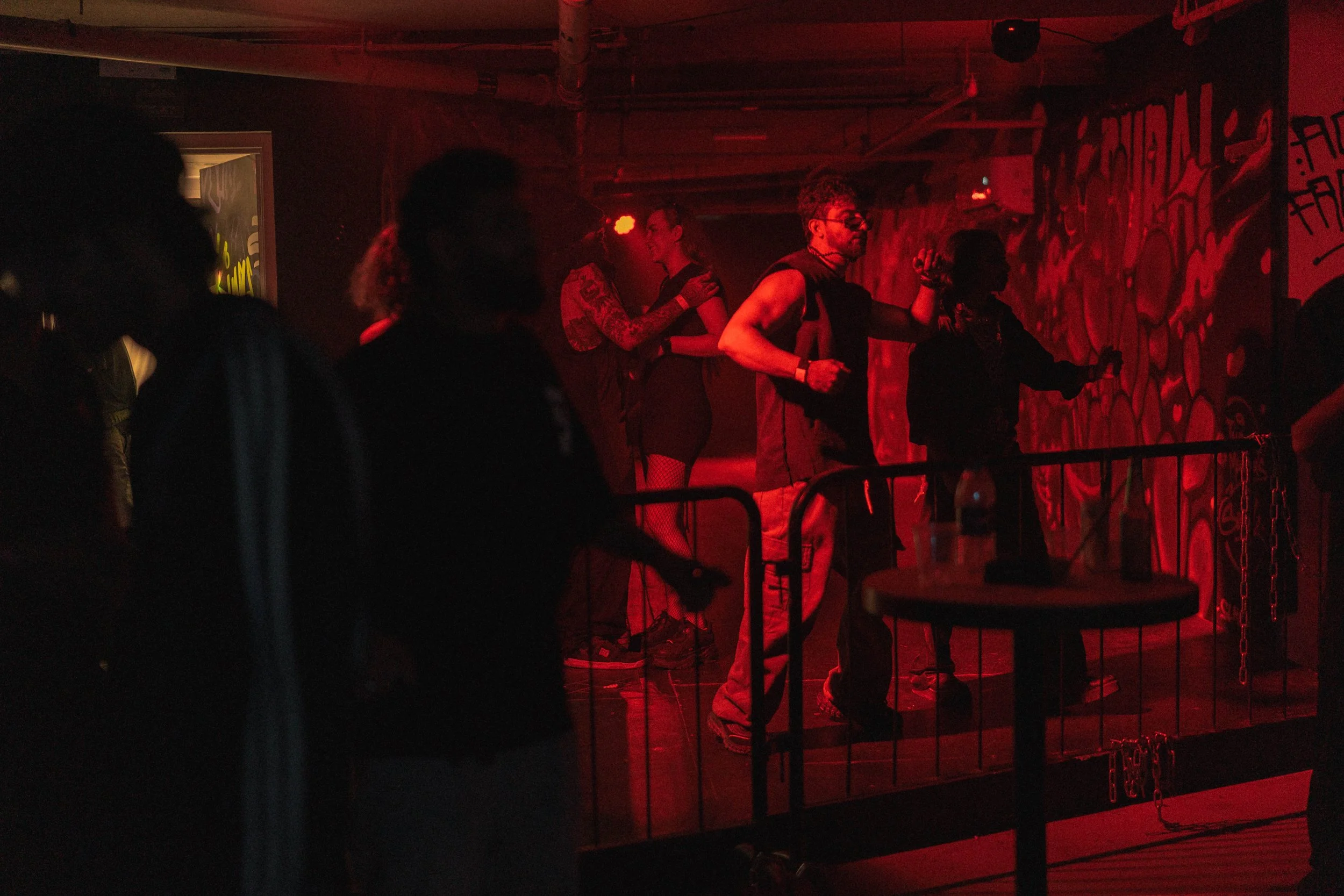 People dancing on a small stage in a dimly lit, red-toned club with graffiti-covered walls.