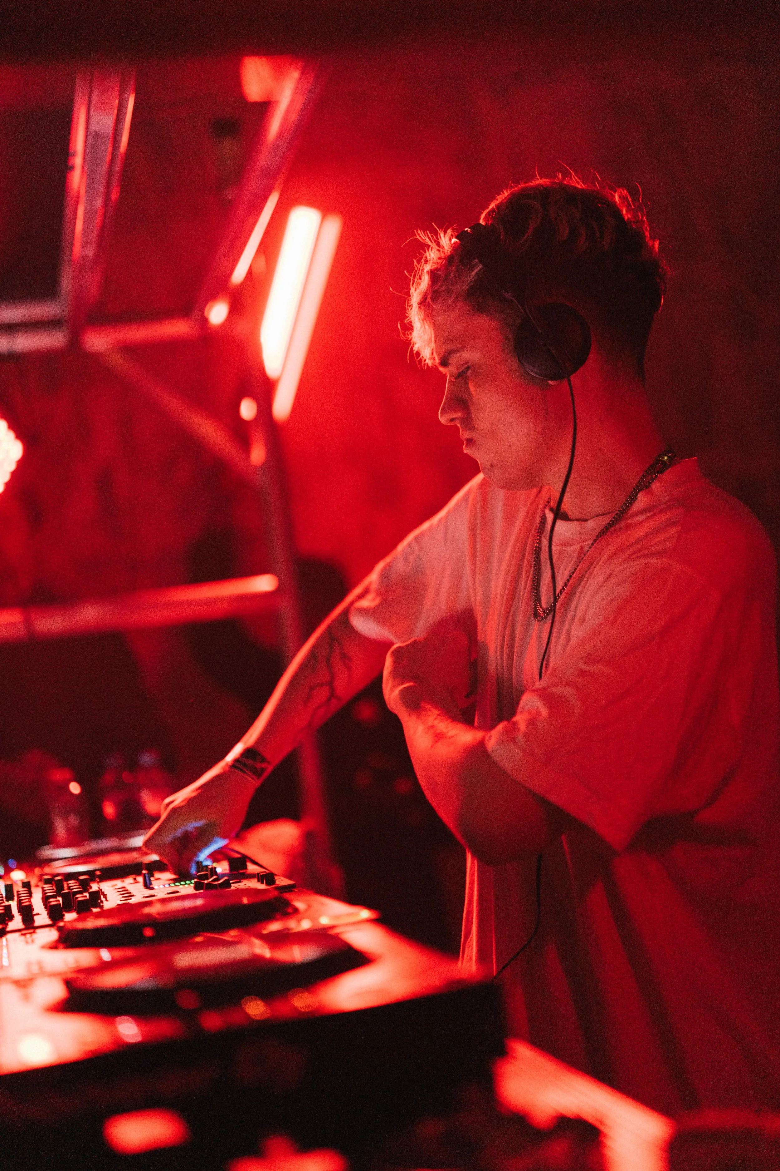 A young male DJ is performing with headphones on, adjusting equipment in a dimly lit setting with red lighting.
