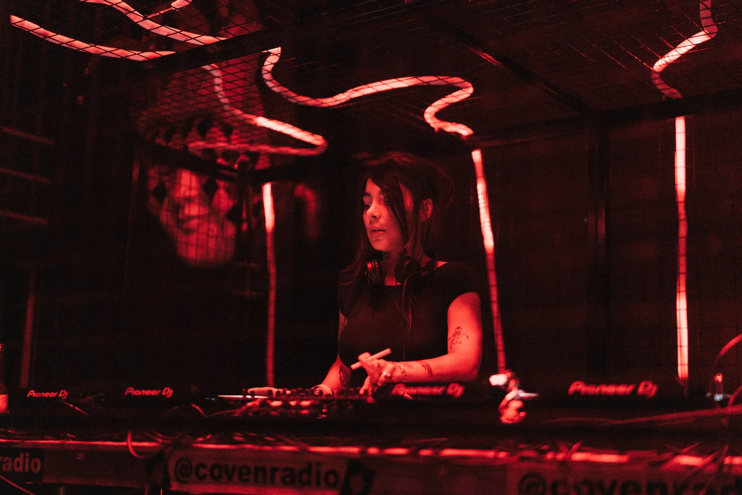 A female DJ with long dark hair and tattoos on her arms, wearing headphones, performing at a club with red lighting, with Pioneer DJ equipment in front of her.