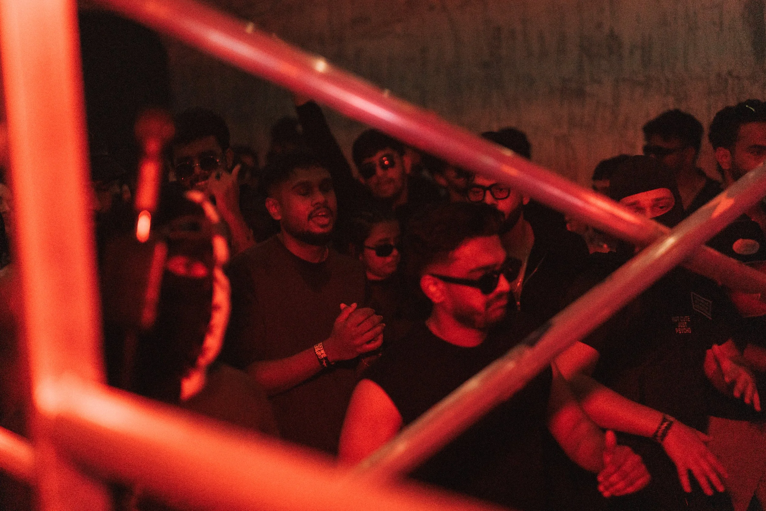 Crowd of people at a nightclub or concert, illuminated by red lighting, some with sunglasses, behind metal bars.