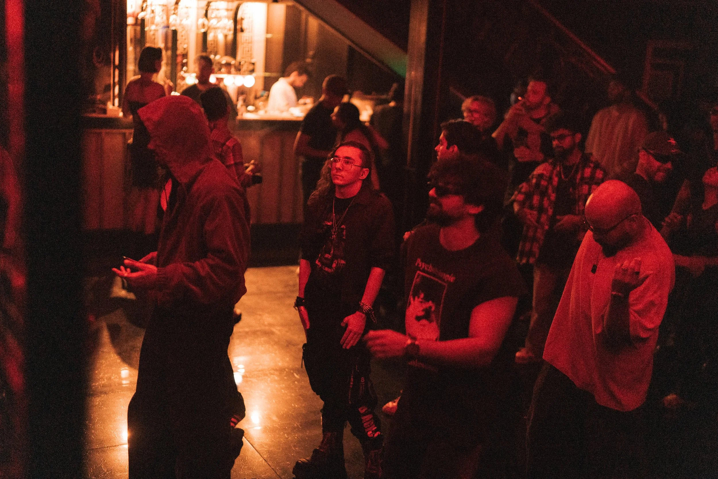 People gathered in a dimly lit indoor space, possibly a music venue or bar, with some standing and looking towards the stage or performance area. There is a bar in the background with people ordering drinks, illuminated by warm lighting.