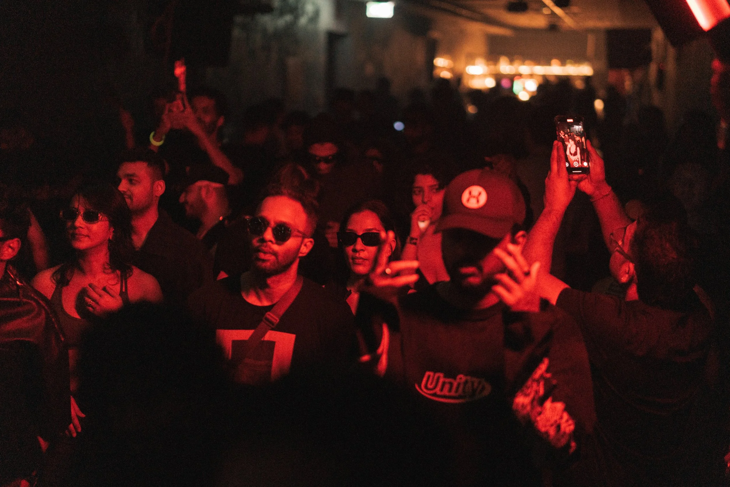 A group of people attending a dimly lit nightclub or concert, some wearing sunglasses. One person is taking a photo or recording with a smartphone. The scene is predominantly illuminated with red lighting.