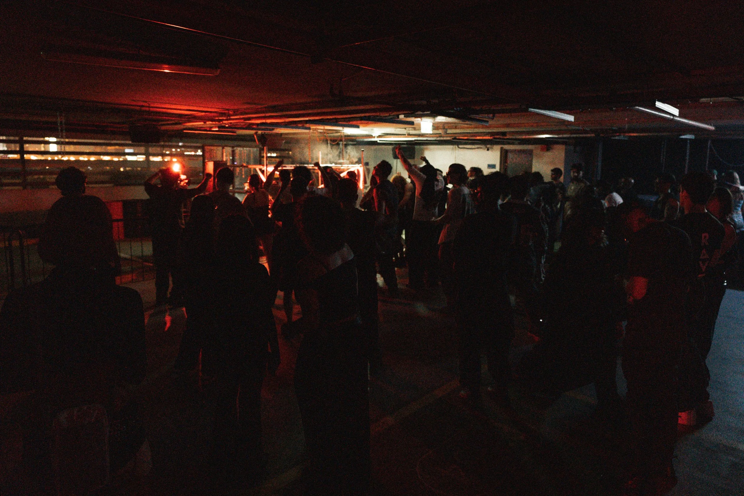Dark indoor parking garage with a group of people gathered, some raising hands, illuminated by red lights.