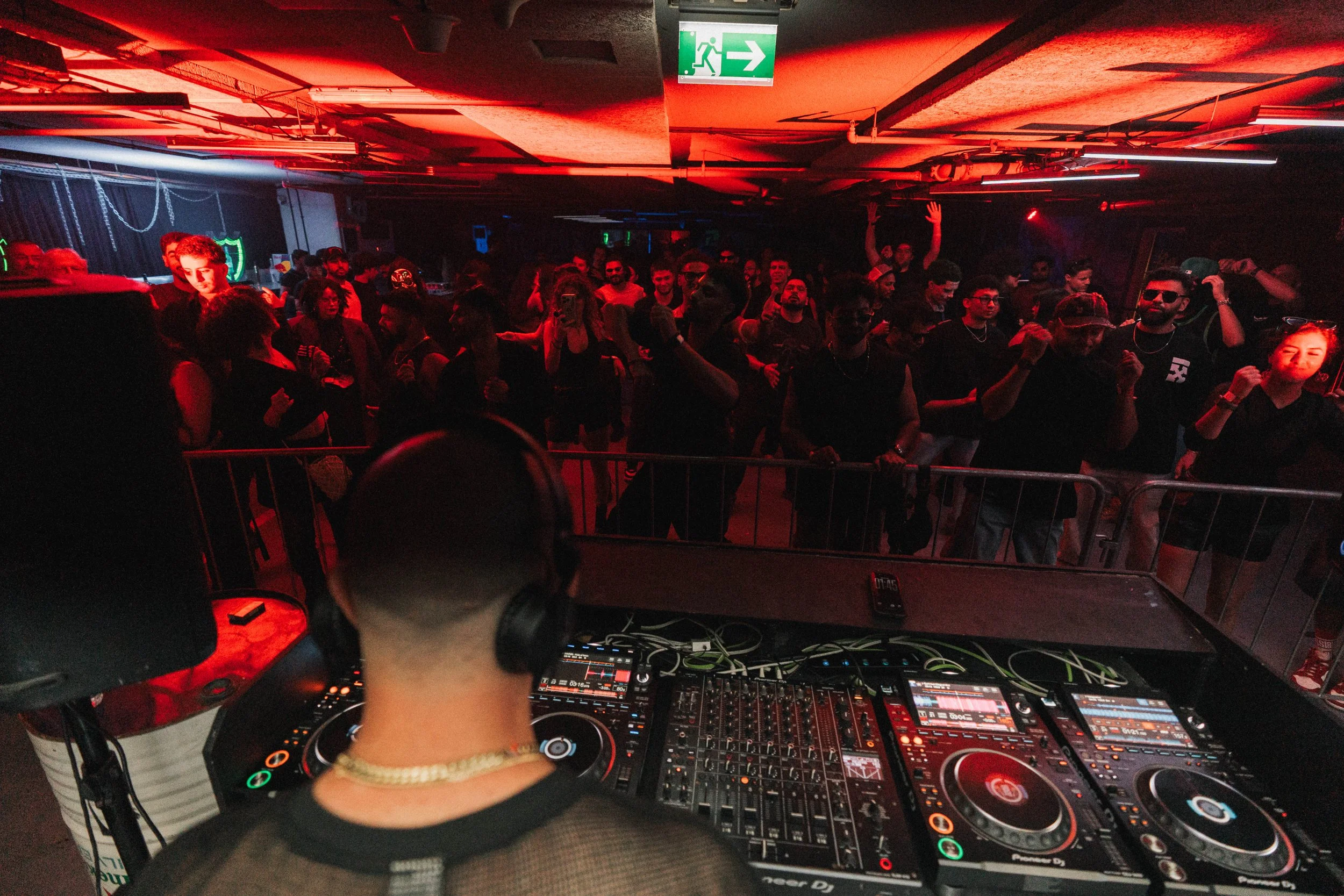 DJ performing at a nightclub with a crowd dancing in front of him, illuminated by red and blue lights, with an emergency exit sign hanging from the ceiling.