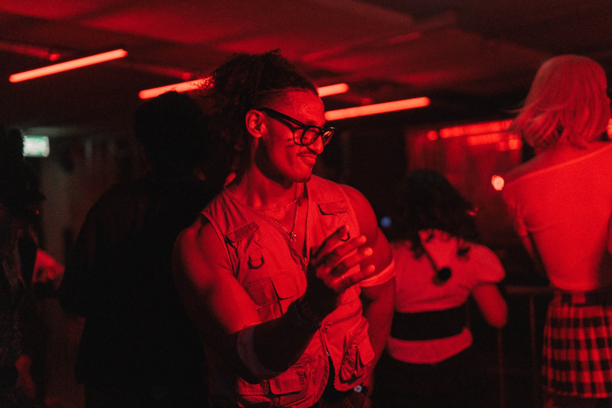 Person dancing at a party or club with red lighting, wearing glasses and a sleeveless vest.