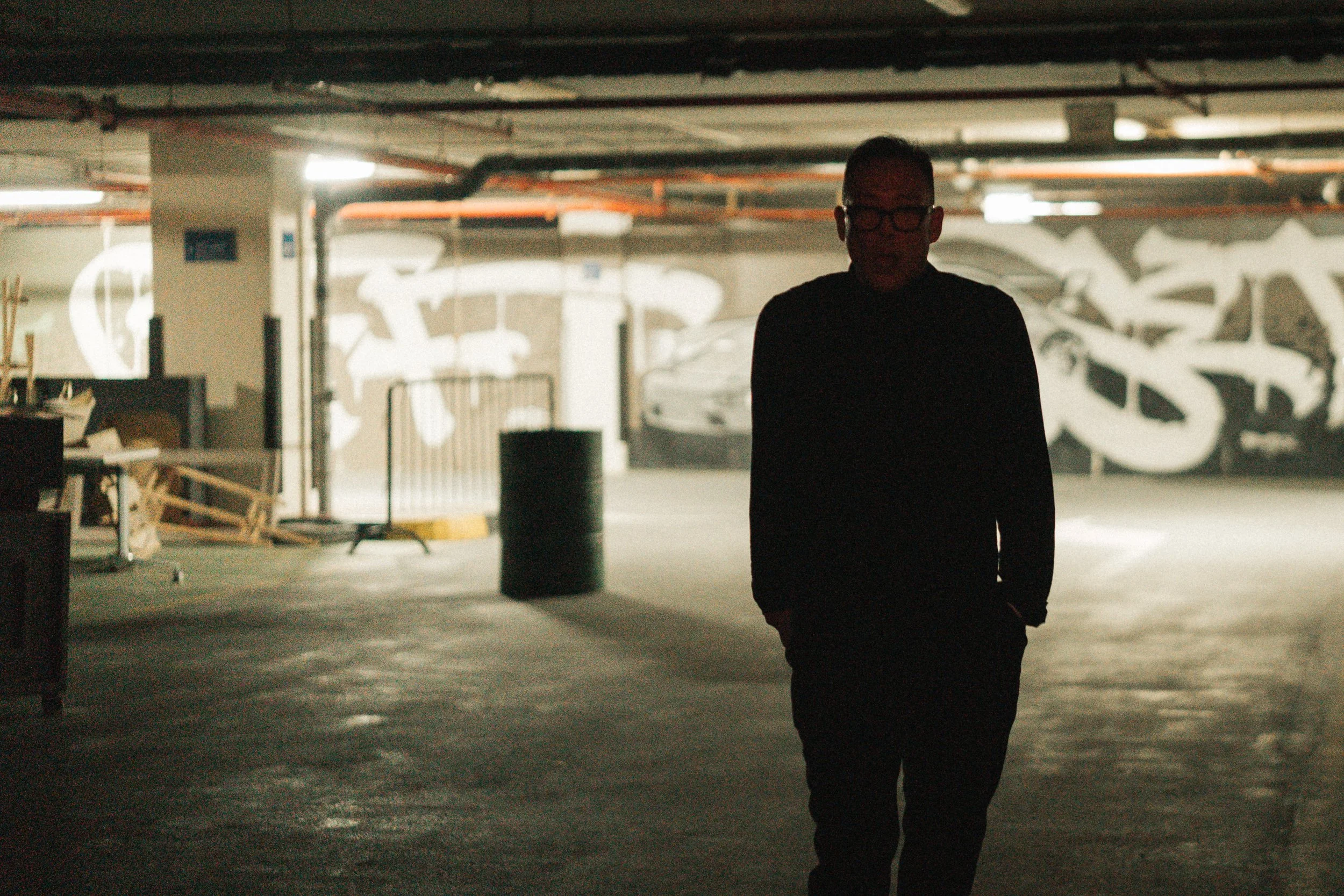 Silhouette of a man with glasses walking in a dimly lit parking garage with graffiti on the wall behind him.