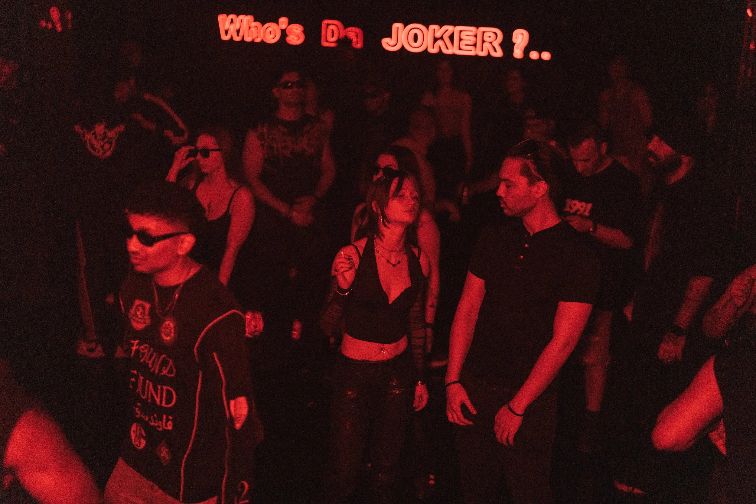 A group of people dancing and socializing in a dark nightclub with red lighting, with a neon sign above that reads 'Who's Da JOKER ?..'