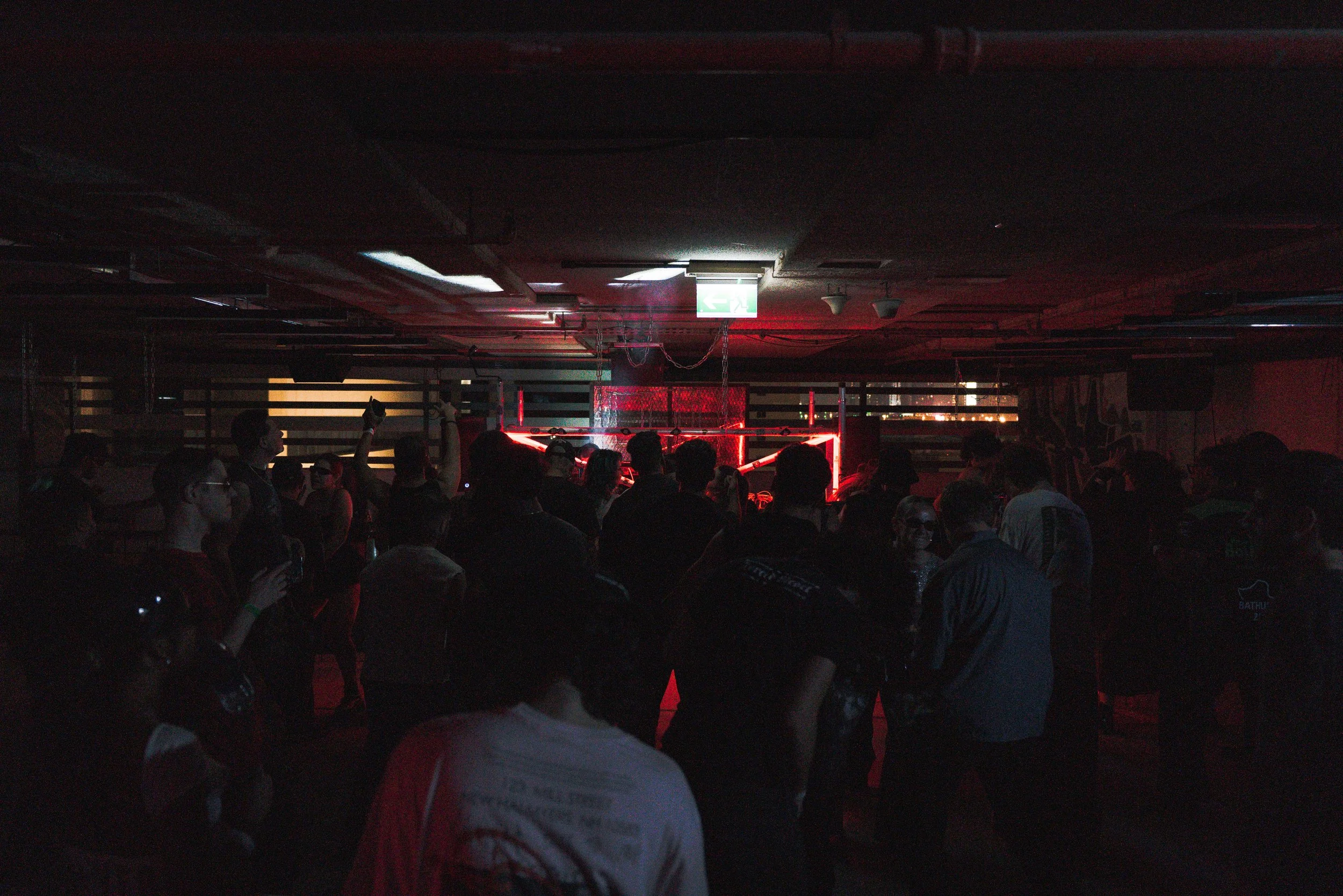 Dimly lit indoor space with a crowd of people standing and socializing, illuminated by red and white lights from stage or DJ setup at the front.