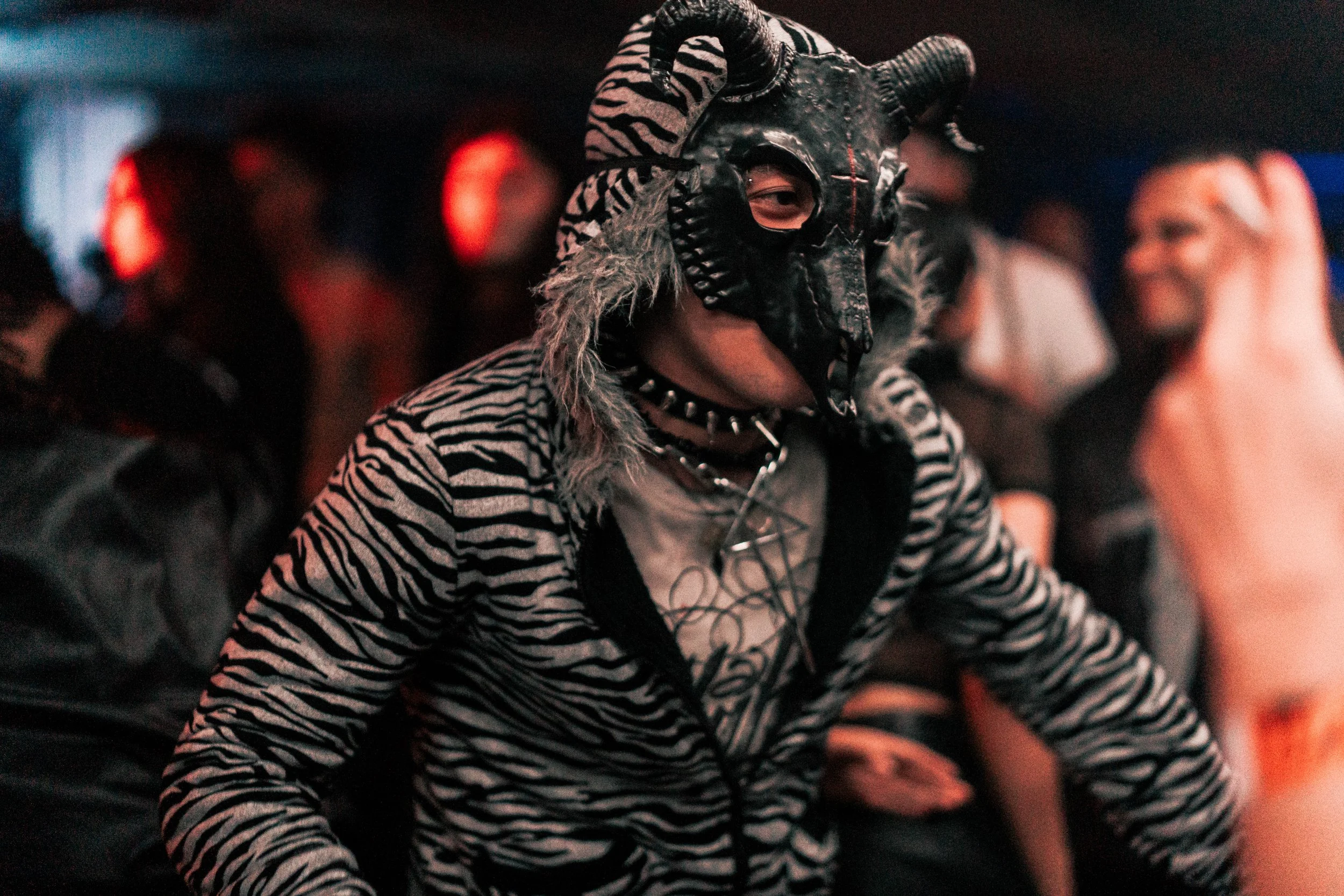 Person wearing a black horned mask, zebra-patterned hoodie, and spiked choker at a party or event.