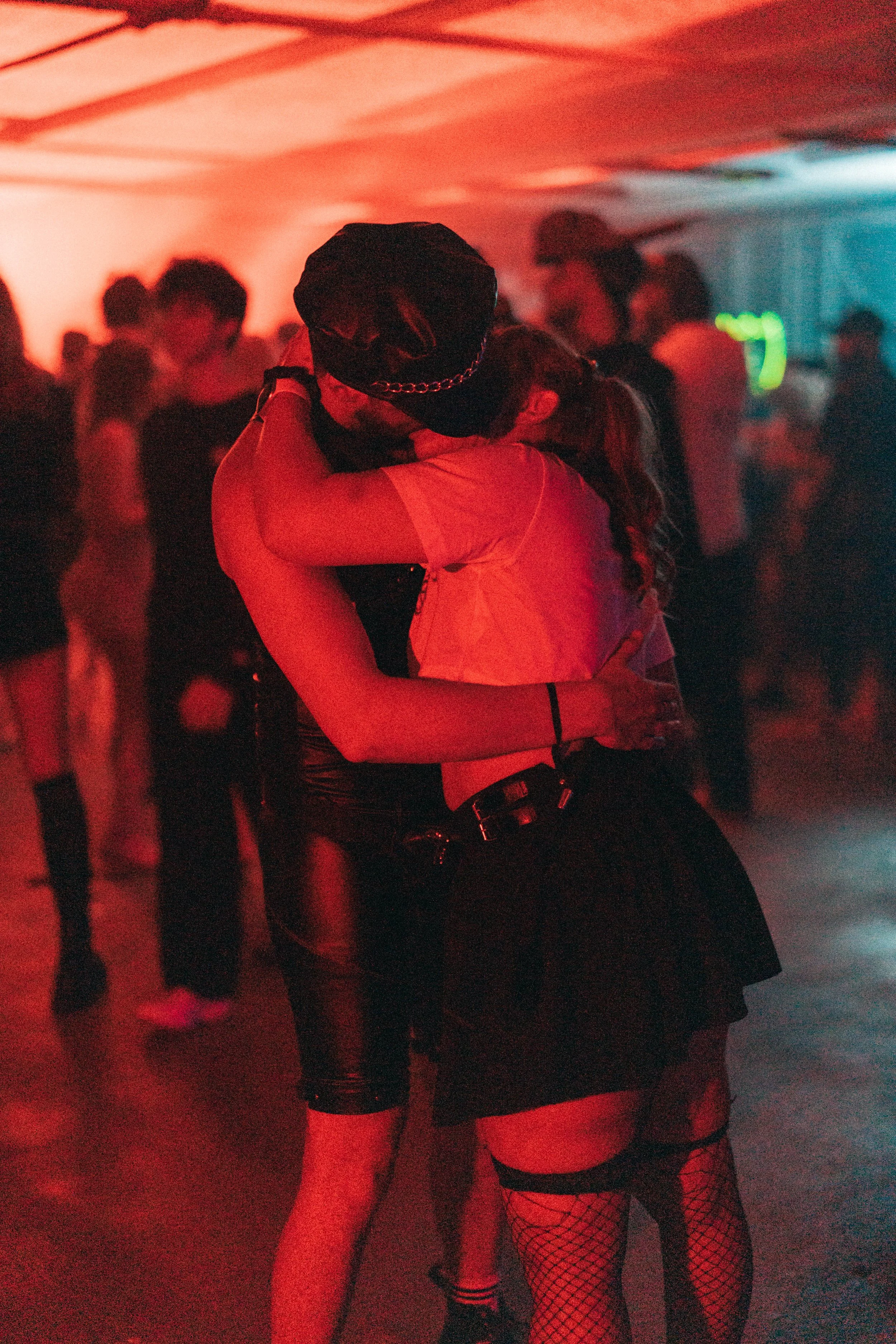 Two women embracing in a dimly lit dance or party setting with red and blue lighting, other people in the background.