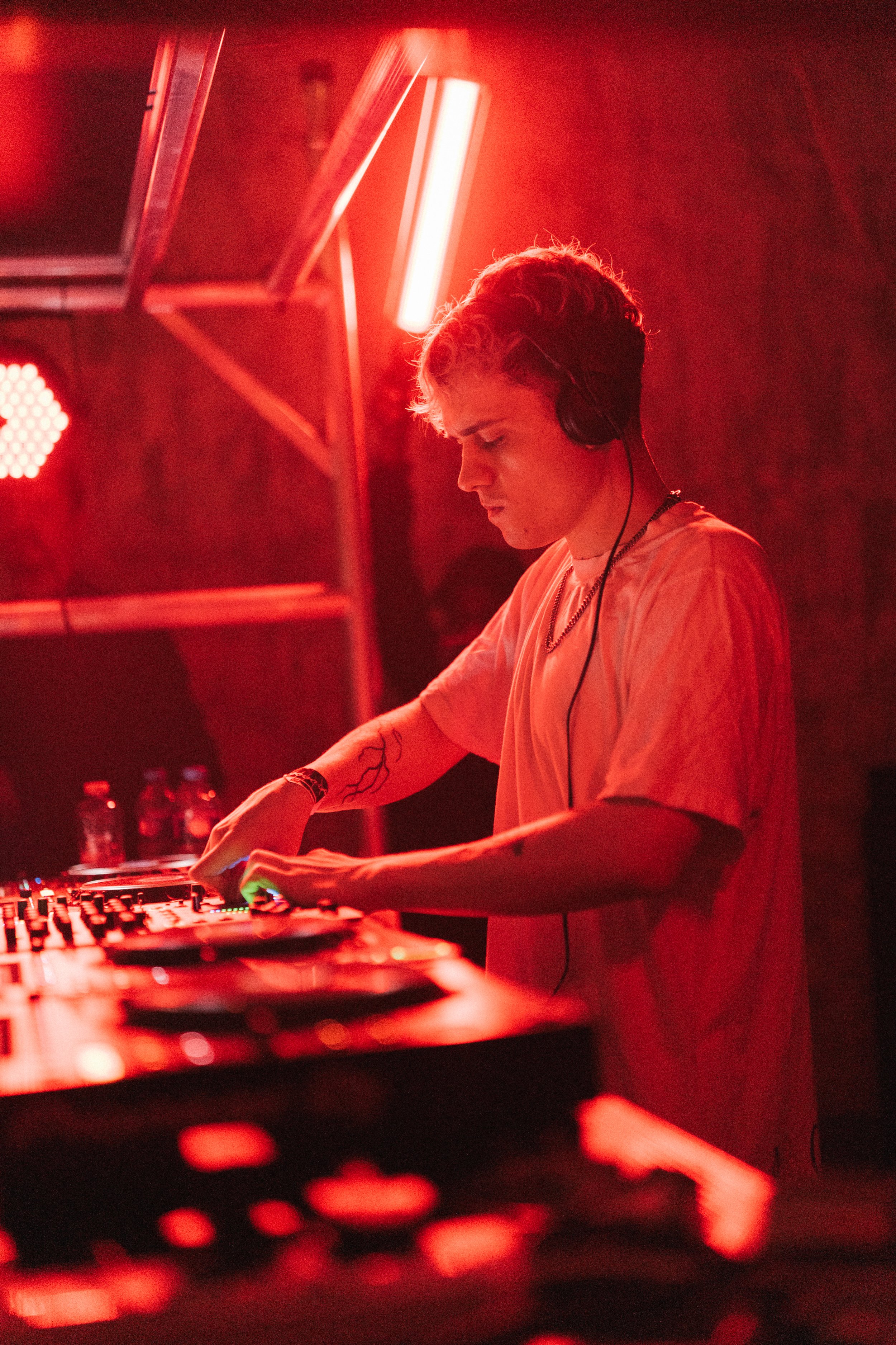 A DJ wearing headphones and a loose t-shirt, mixing music at a DJ booth illuminated in red light.