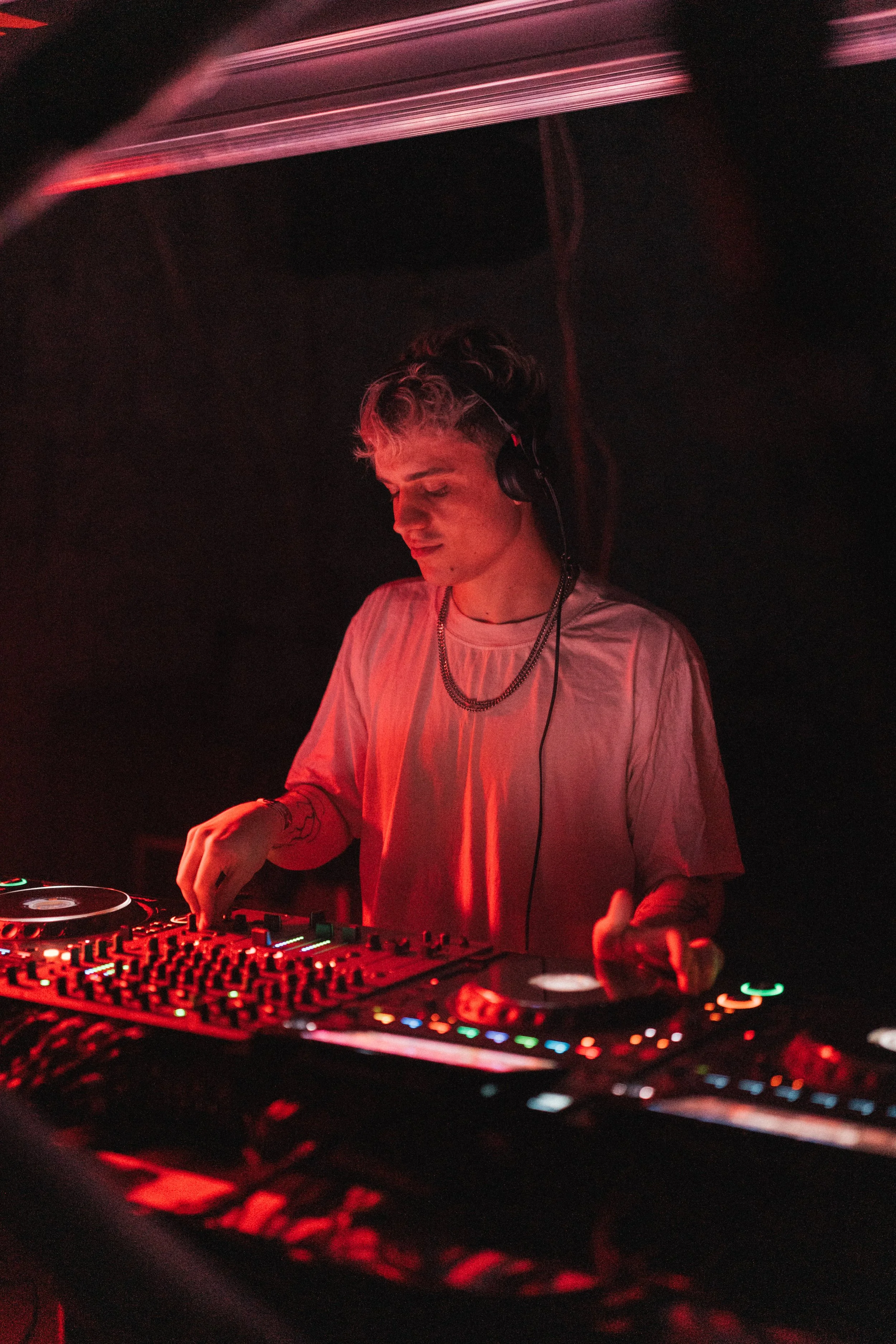 A young male DJ with short, wavy blonde hair and tattoos on his arms, wearing headphones and a silver chain necklace, performing on a DJ deck in a dark environment with red lighting.