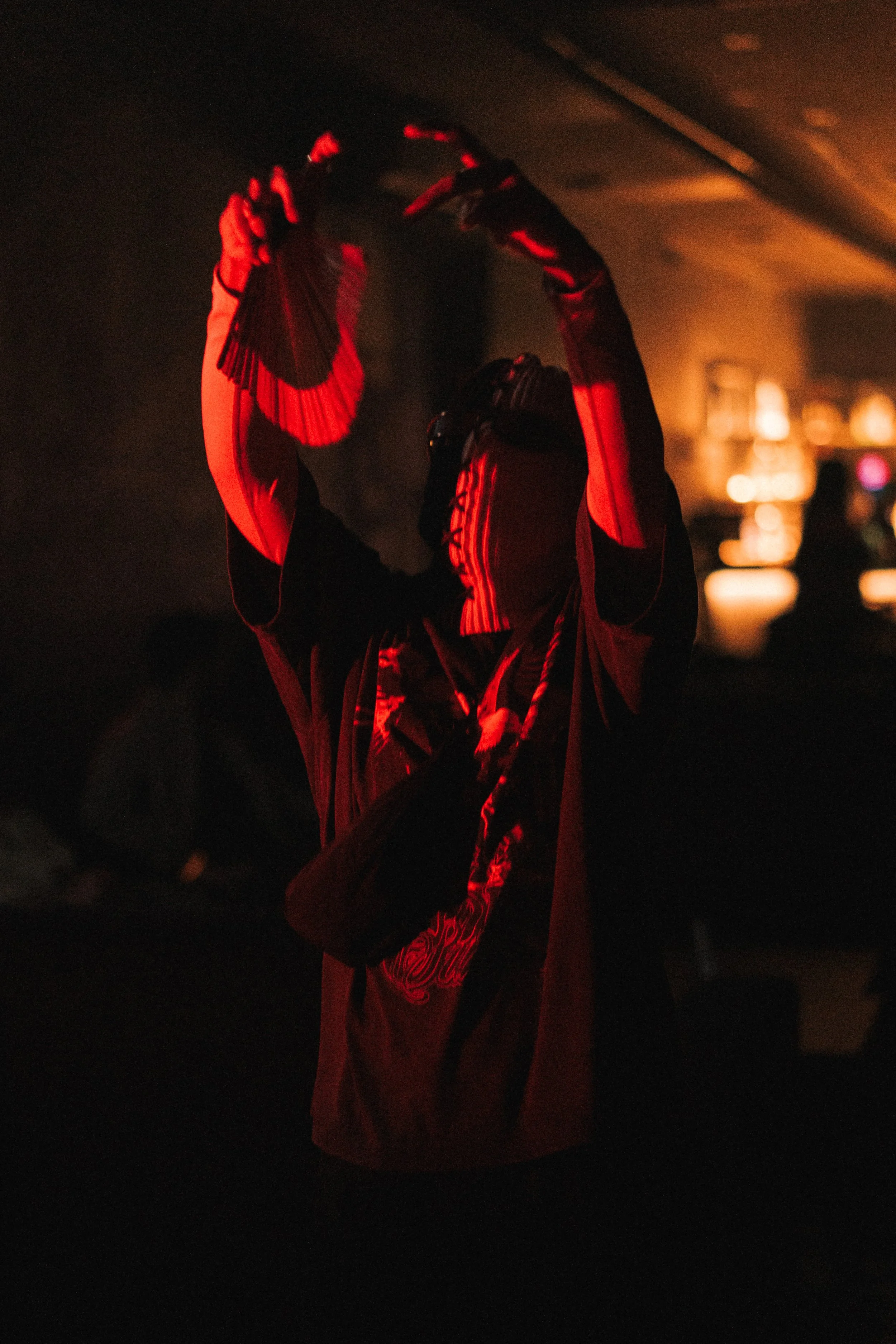 A person dancing in a dark room illuminated by red light, with hands raised above their head, wearing a black shirt with red patterns.