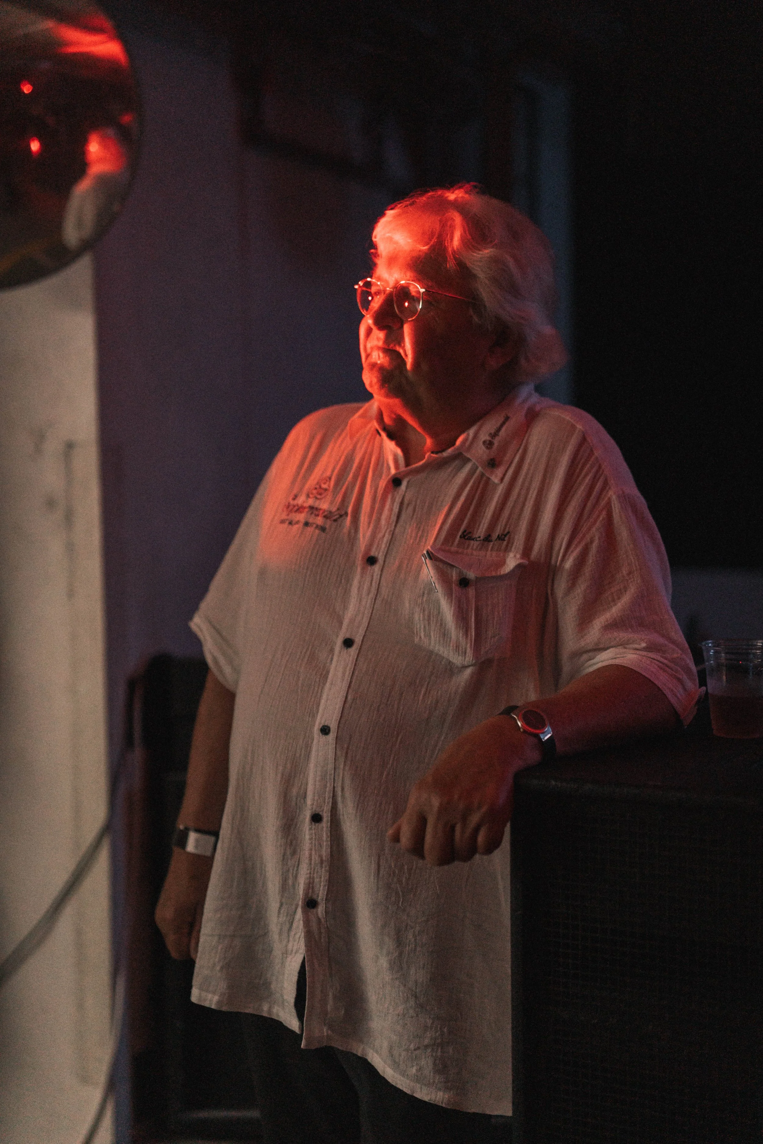 Older man with glasses and white hair wearing a light-colored, striped button-up shirt, standing in low lighting with a red hue, leaning on a surface with a drink, in a dark environment.