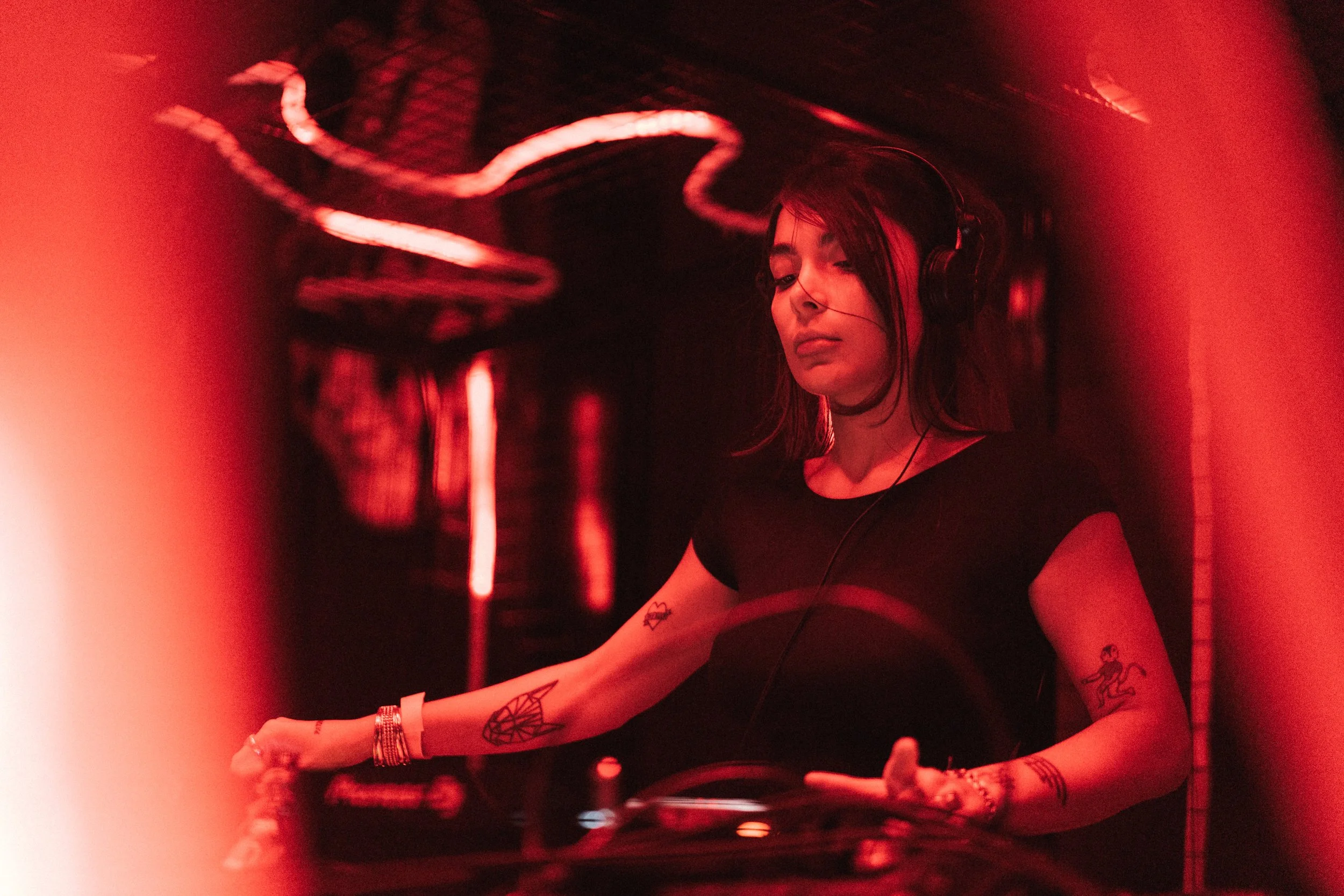 A young woman DJ wearing headphones and a black shirt, with tattoos on her arms, mixing music in a dimly lit, red-toned setting.