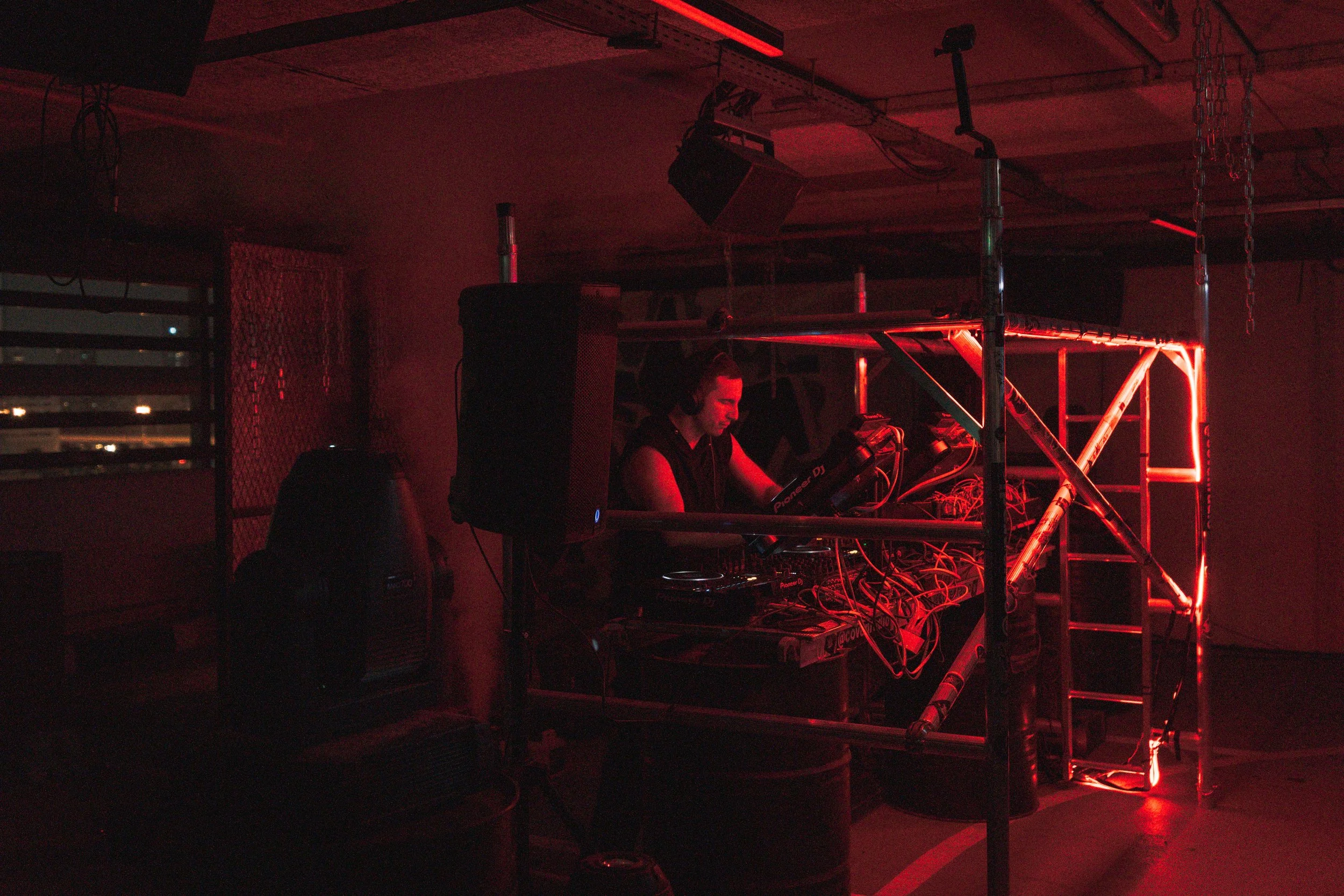 A DJ performing at a nightclub with red neon lighting, surrounded by sound equipment and scaffolding.