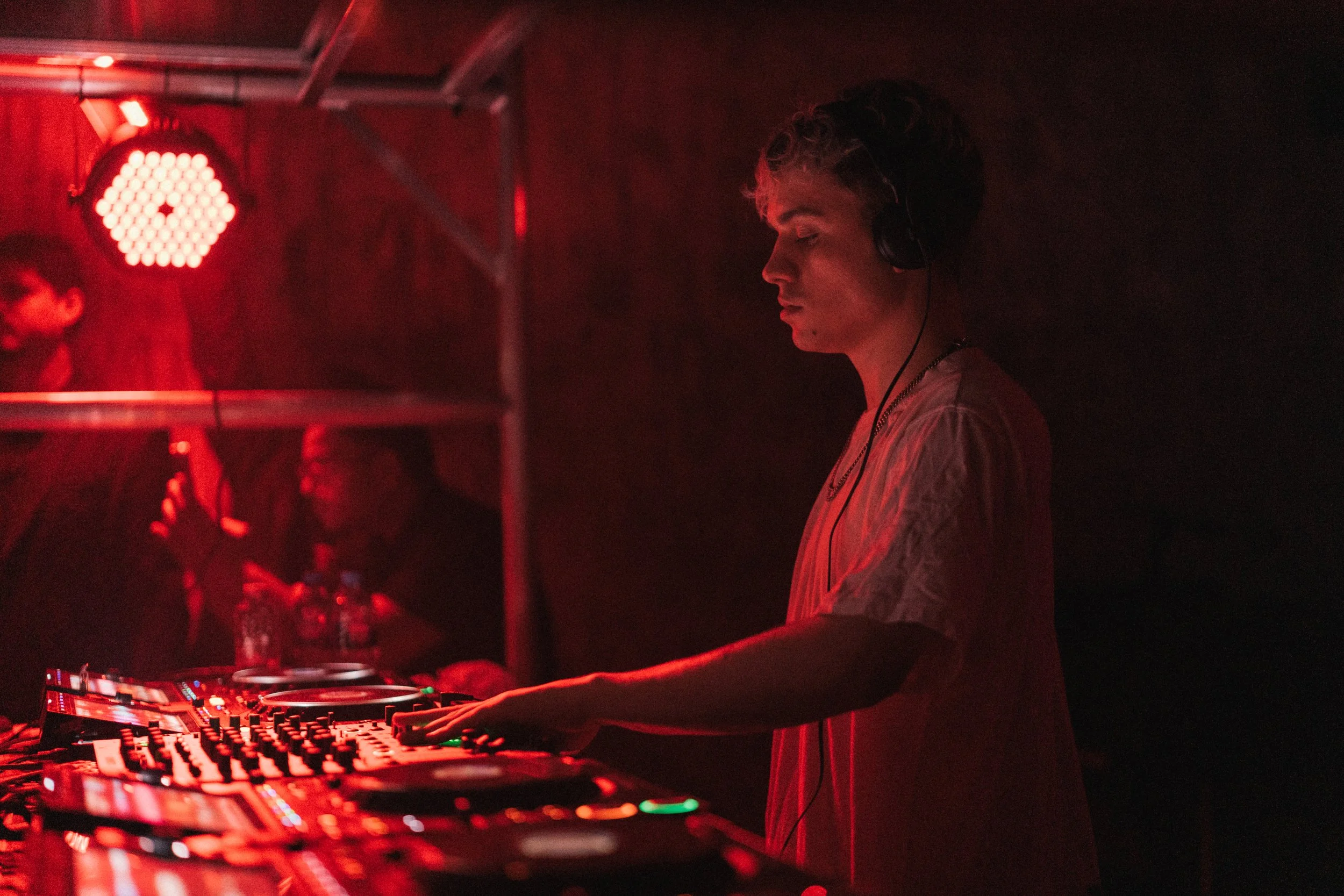 A male DJ wearing headphones is playing music behind a DJ mixing console in a dimly lit, red-hued environment.