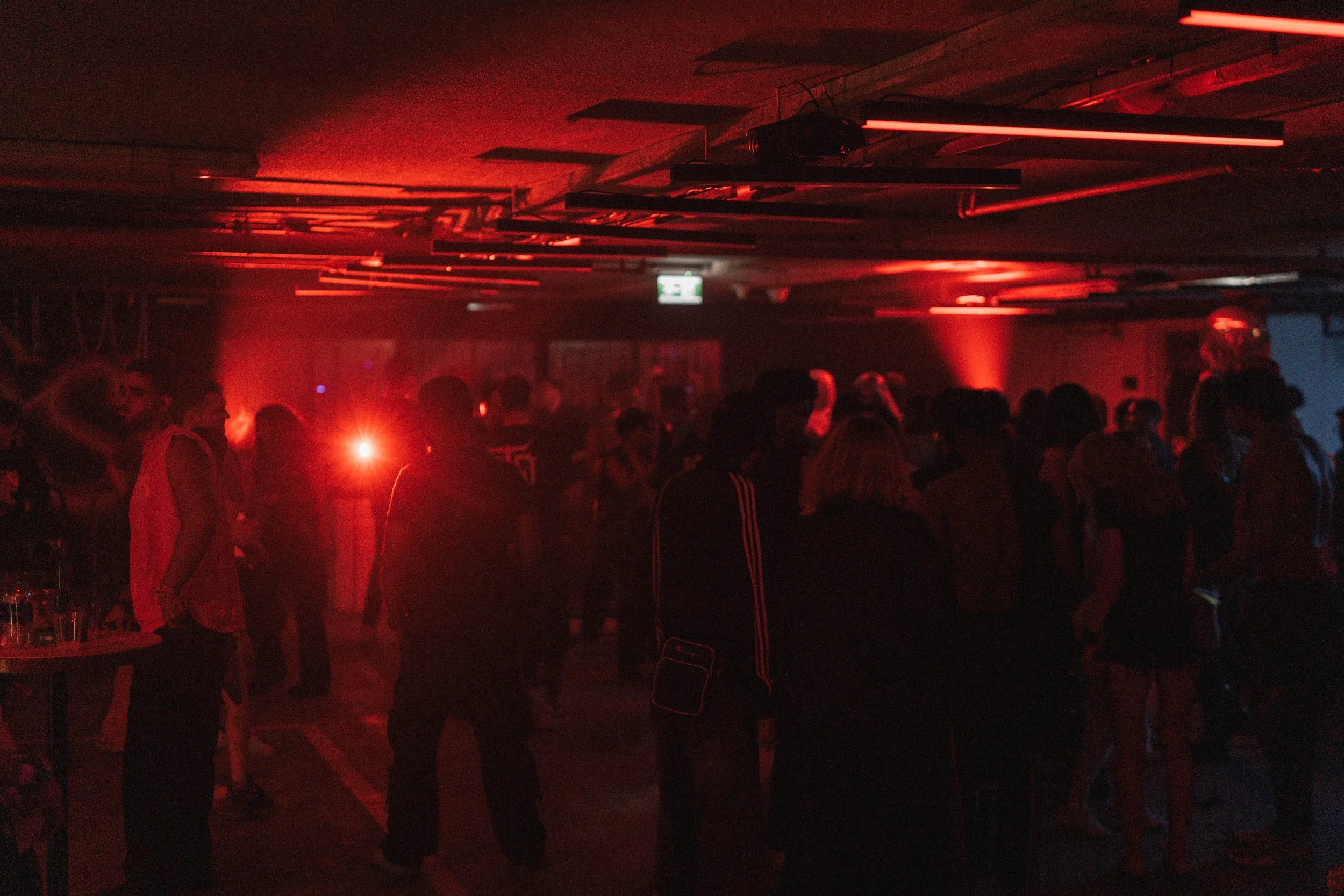 People dancing and socializing in a dark, red-lit indoor setting, possibly a nightclub or party.