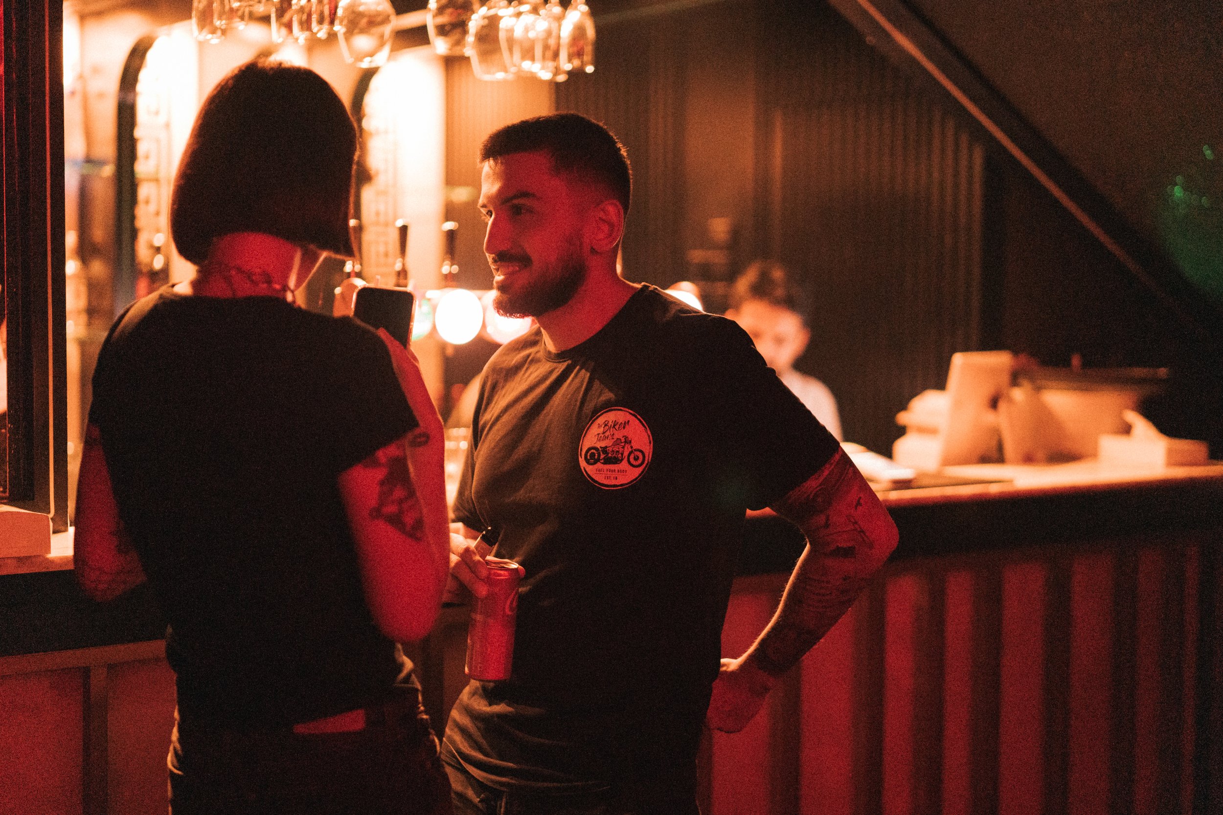 Two people are talking at a bar, one holding a drink and the other with a phone. The bar has warm lighting and a bartender is working in the background.