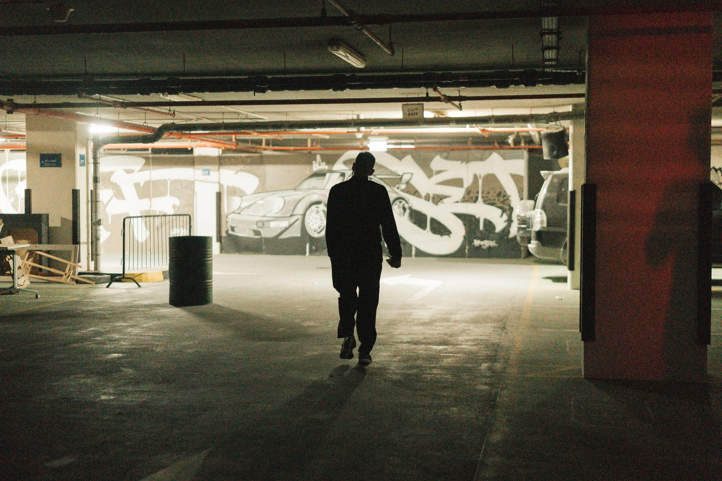 A person walking in a dimly lit underground parking garage with graffiti art of a car on the wall in the background.