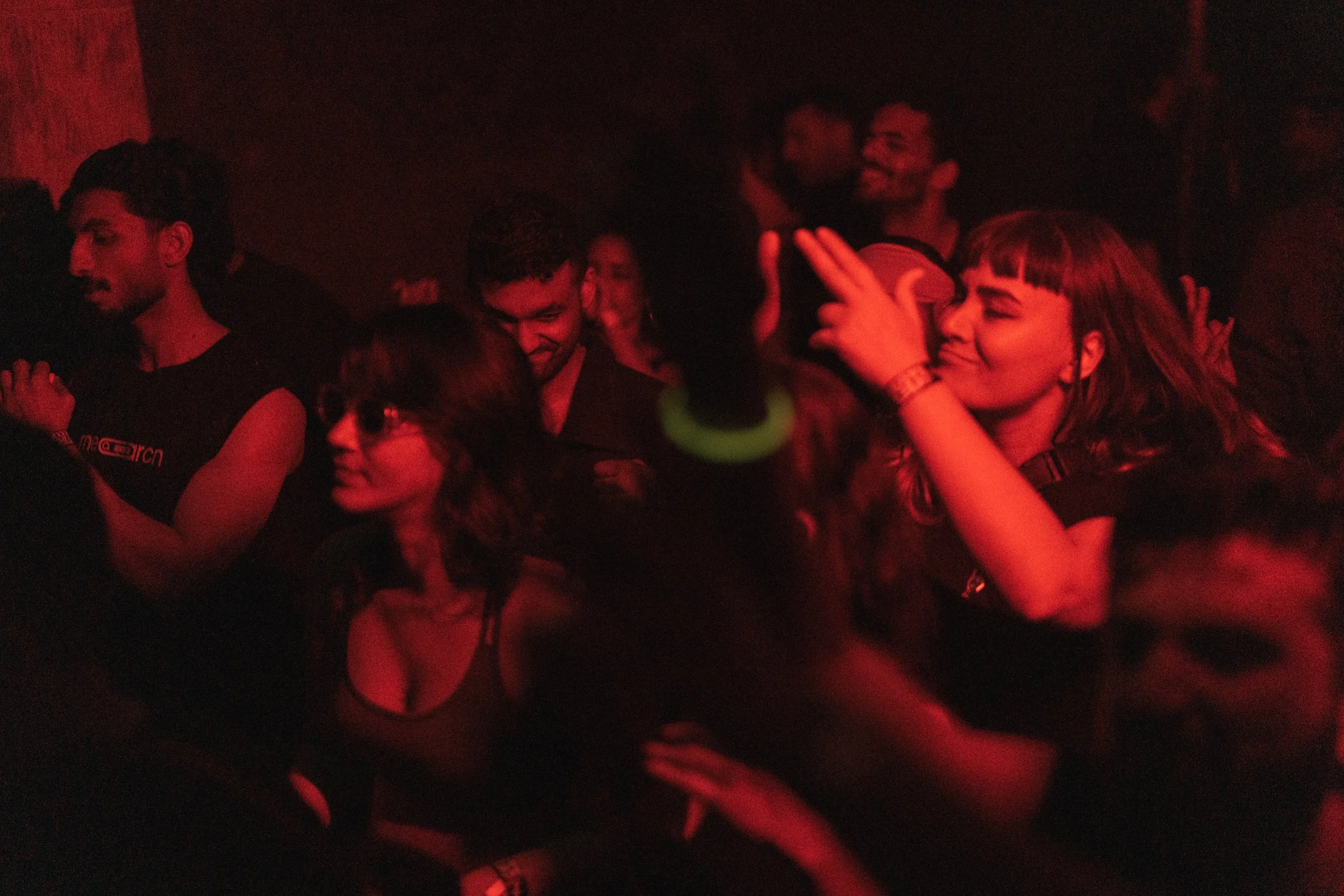 People dancing and enjoying music at a dark, red-lit concert or party, with some individuals smiling and raising their hands.