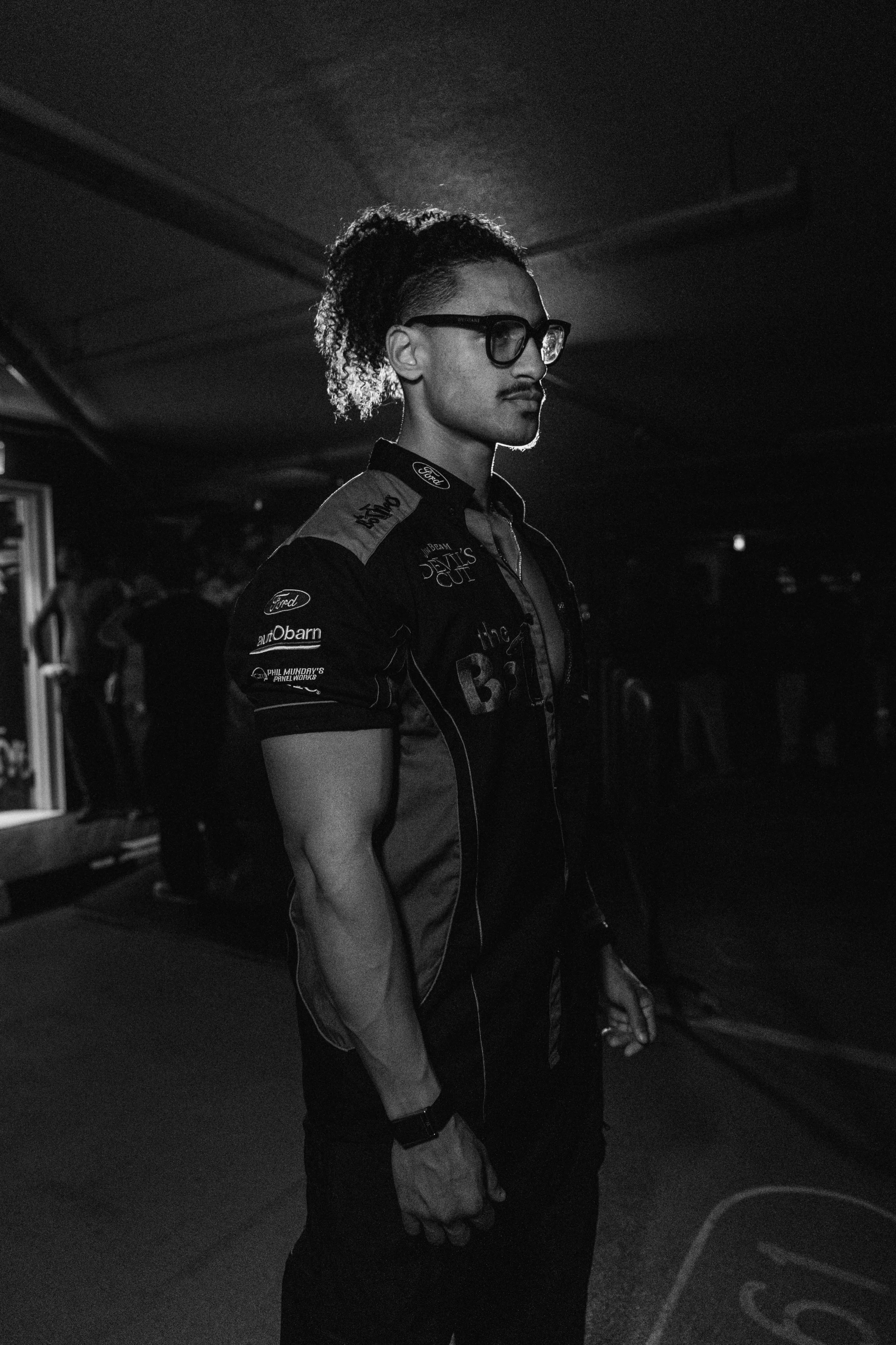 A man with curly hair, glasses, and a mustache wearing a racing suit with logos stands in a dimly lit area.