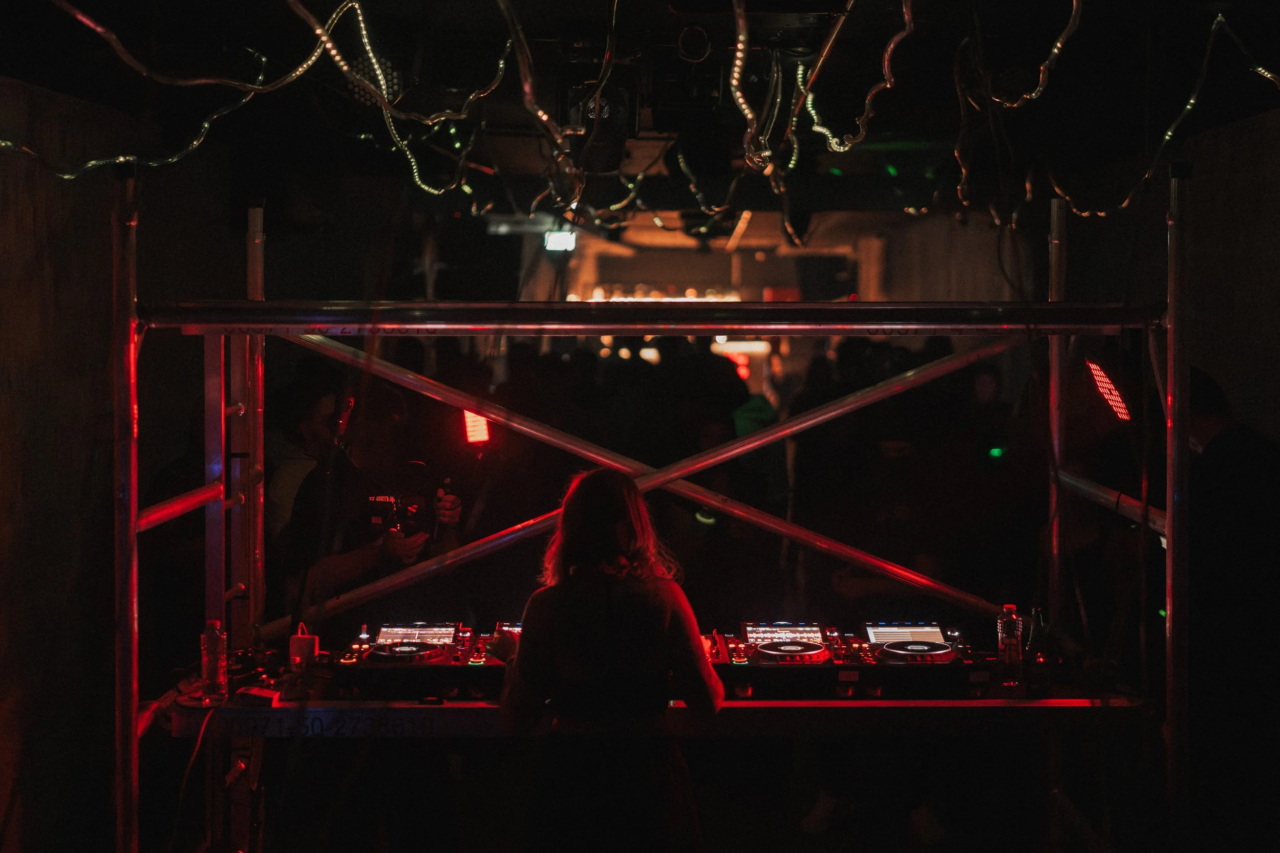 A DJ performing at a nightclub with a dark background, illuminated by red and green lights, surrounded by speakers and electronic equipment.