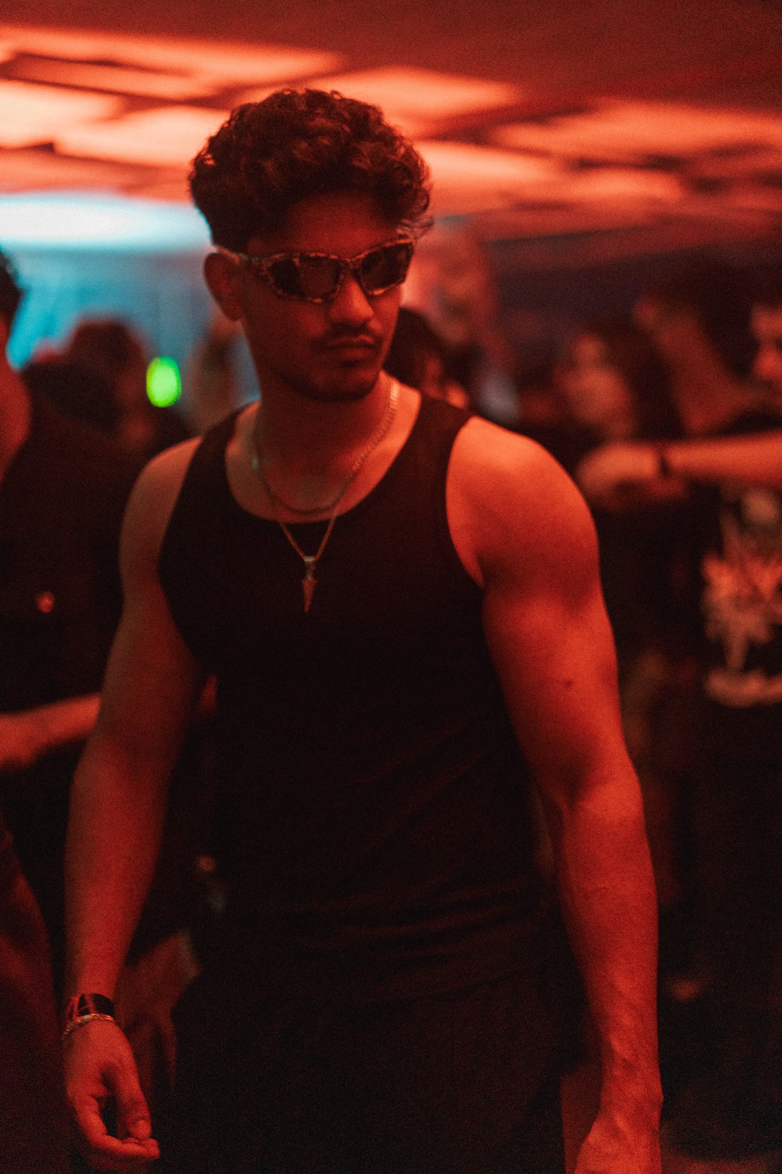 A young man with styled hair wearing sunglasses, a black sleeveless shirt, a necklace, and a watch, standing in a dimly lit nightclub with red and blue lighting, surrounded by people.